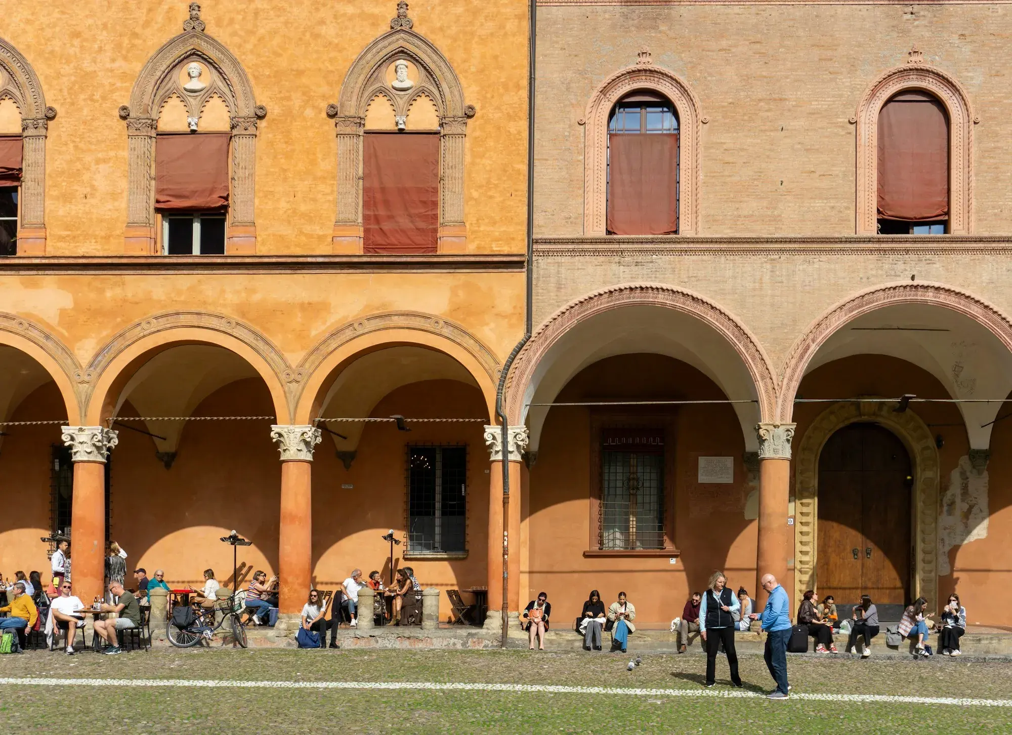 Arcaded porticoes with red and ochre columns in central Bologna, with people sitting and walking beneath them