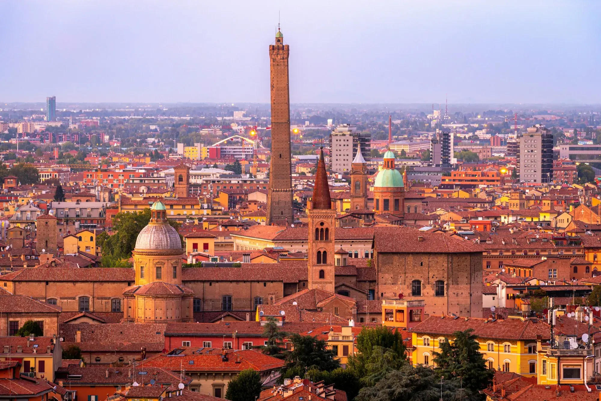 Bologna's red and ochre rooftops seen from the hills at dusk, with the tall Asinelli tower, church domes and bell towers rising above the city