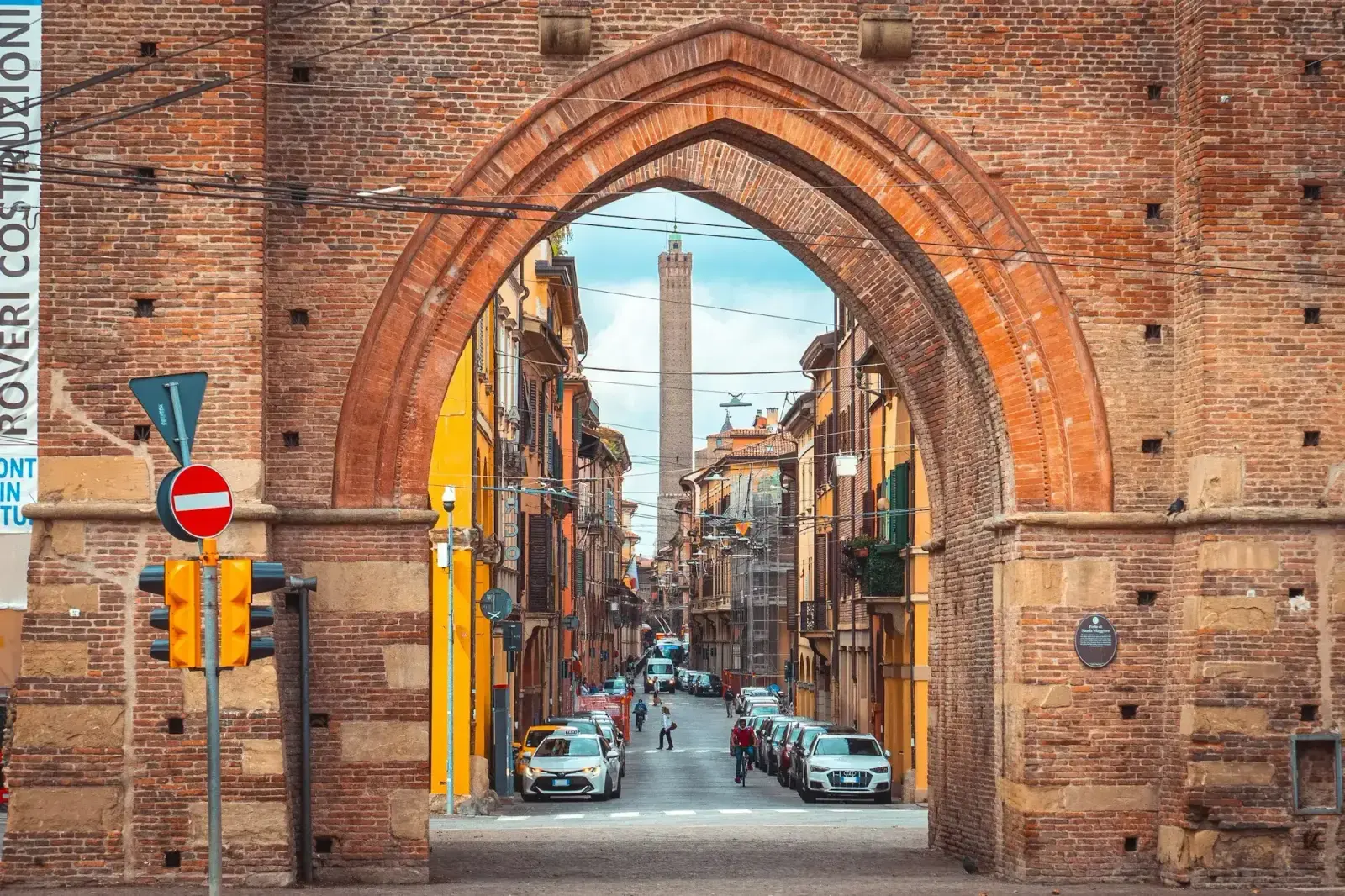 Strada Maggiore in Bologna seen through the brick arch of Porta Maggiore, with arcaded buildings and a tall medieval tower at the far end