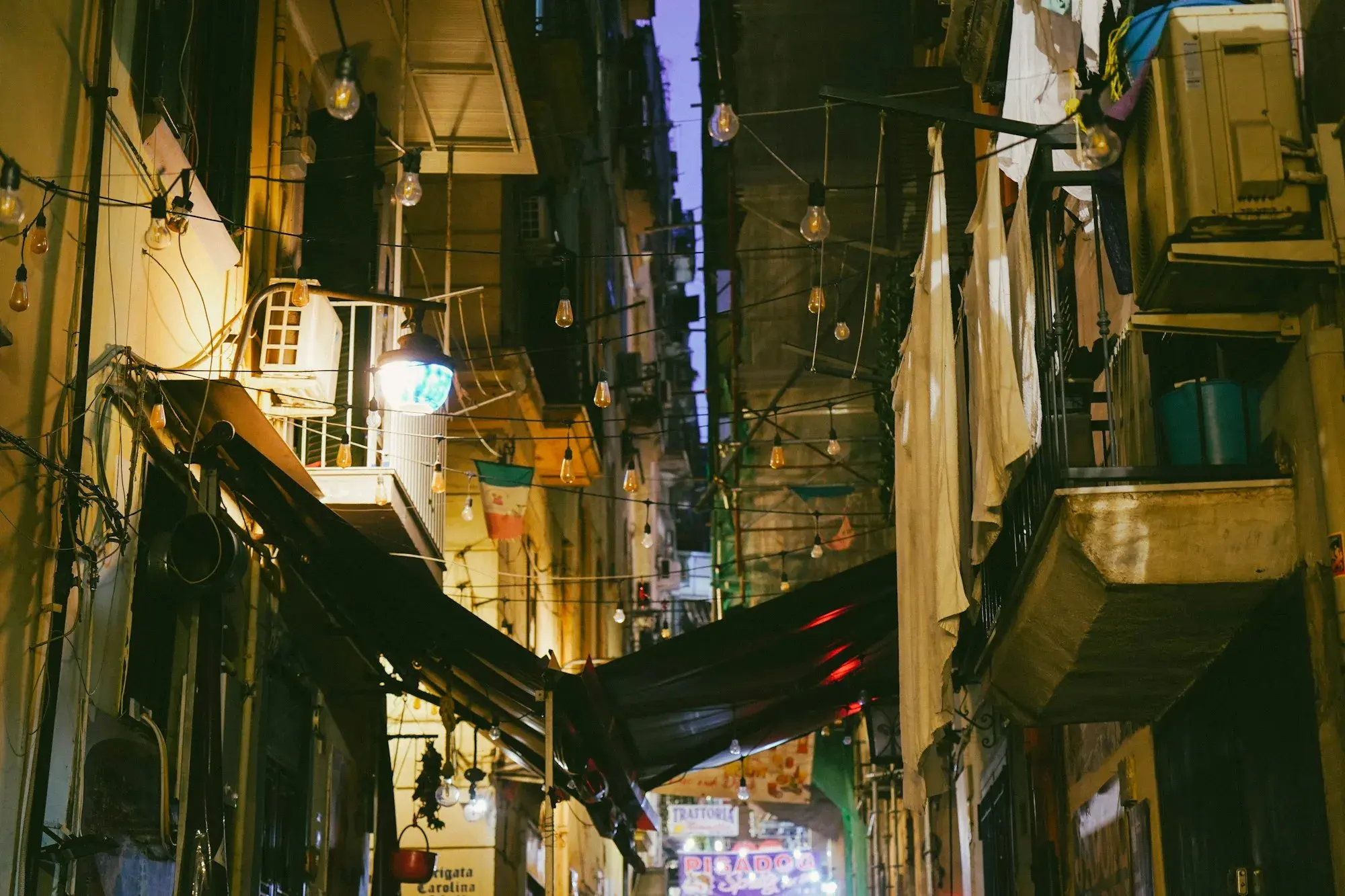 A narrow street in central Naples at night with string lights, balconies and restaurant signs