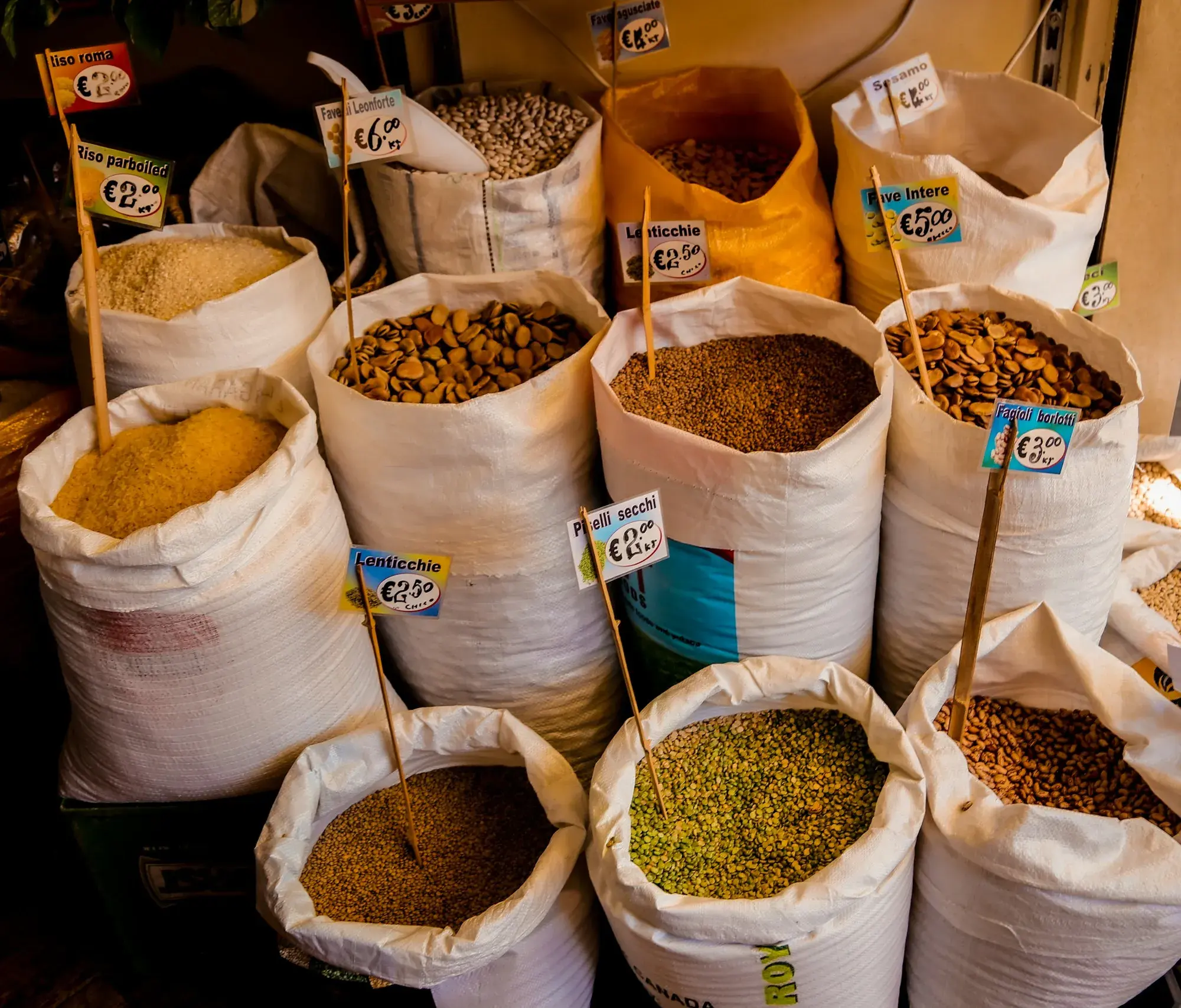 Open sacks of grains, pulses and dried legumes with handwritten price labels at a market stall in the Albergheria district