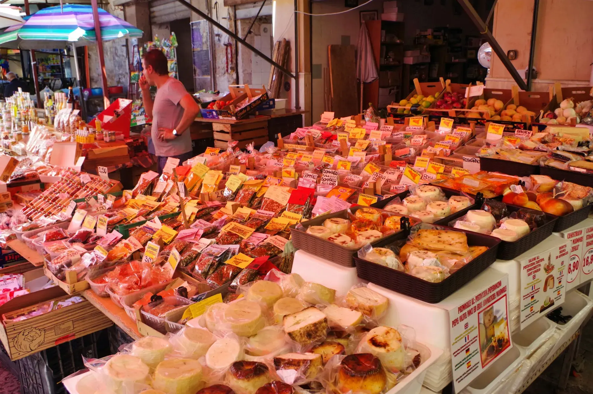 A stall at Ballarò market in Palermo piled with cheeses, cured meats and packaged foods with price labels, and a stallholder behind