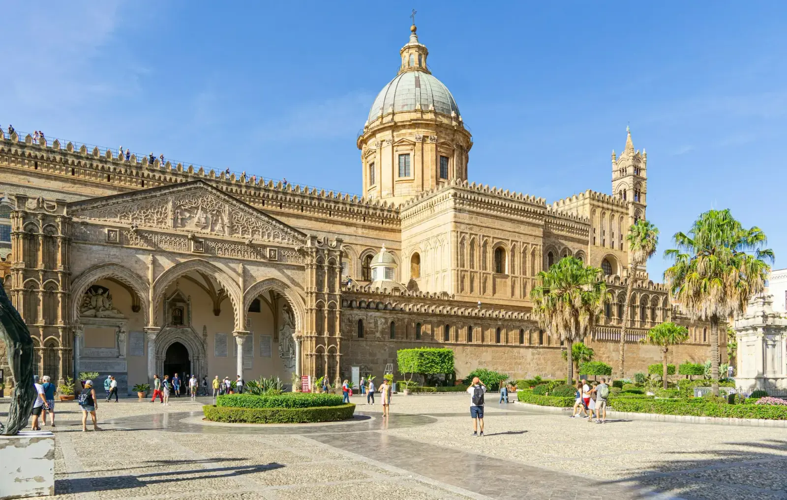 Palermo Cathedral in sunlight, with its Gothic towers, dome and long side portico, and palm trees in the garden in front