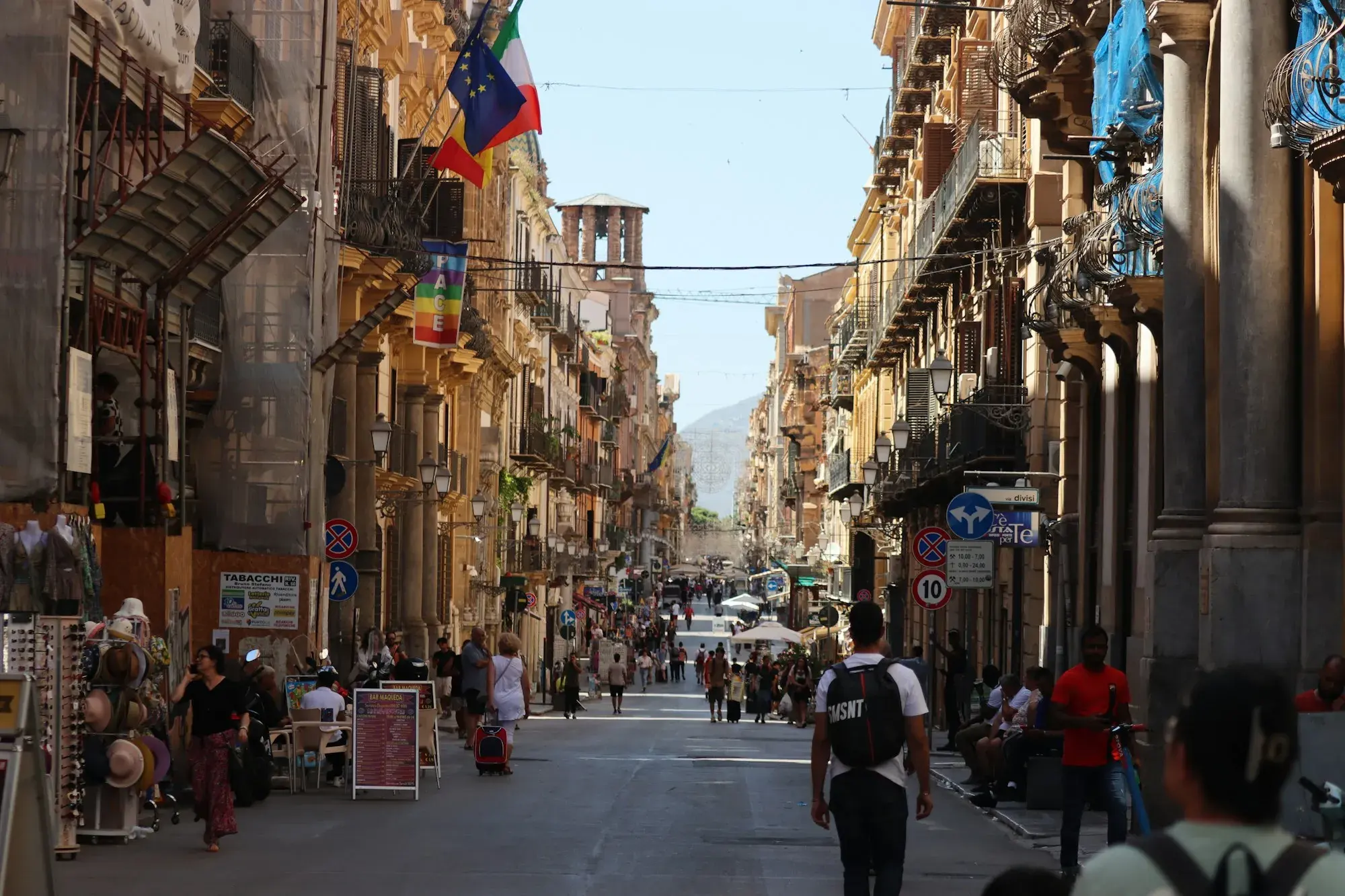 A pedestrian street in central Palermo on a summer day, lined with historic buildings with balconies, with people walking