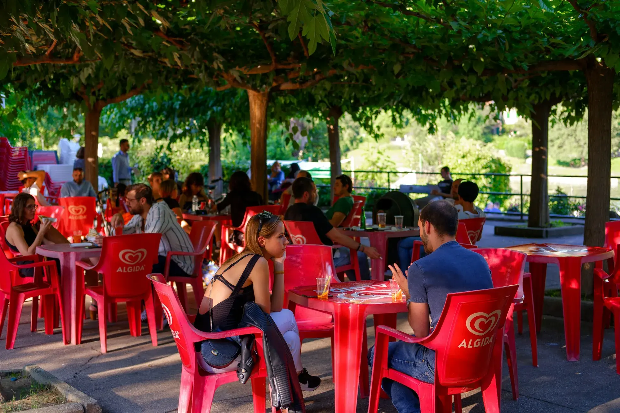 People sitting at red tables at an outdoor café under the trees in the Parco del Valentino, Turin