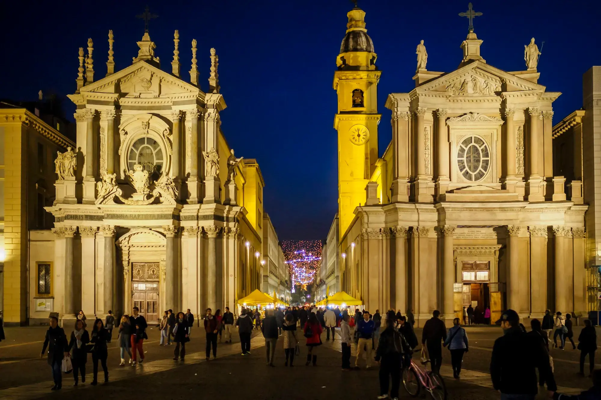 Piazza San Carlo in Turin at dusk, with the twin Baroque churches lit up and people crossing the square
