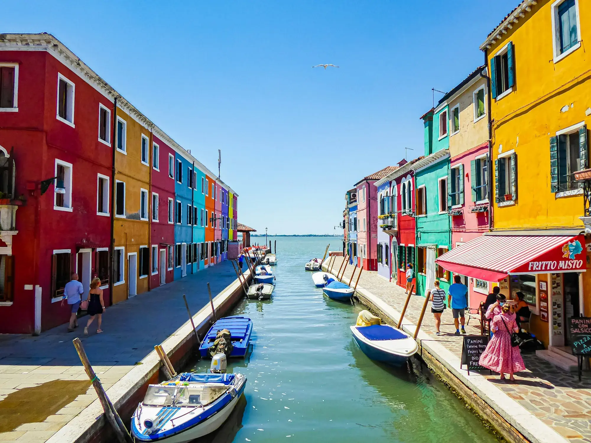 Brightly painted houses in red, orange and blue along a canal on the island of Burano, with small boats moored in the water