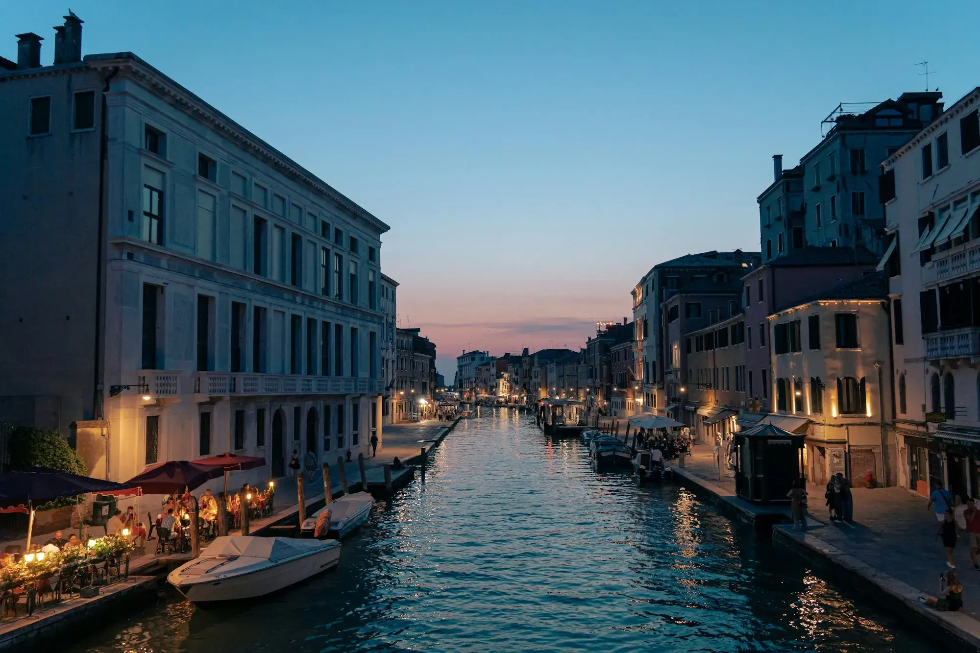 A wide canal in Venice at dusk, with restaurant tables and lights along the waterside path and moored boats