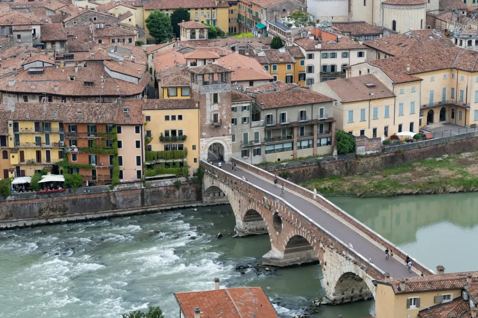 The Ponte Pietra, a stone and brick arched bridge over the fast-flowing River Adige in Verona, with pastel houses and red roofs behind