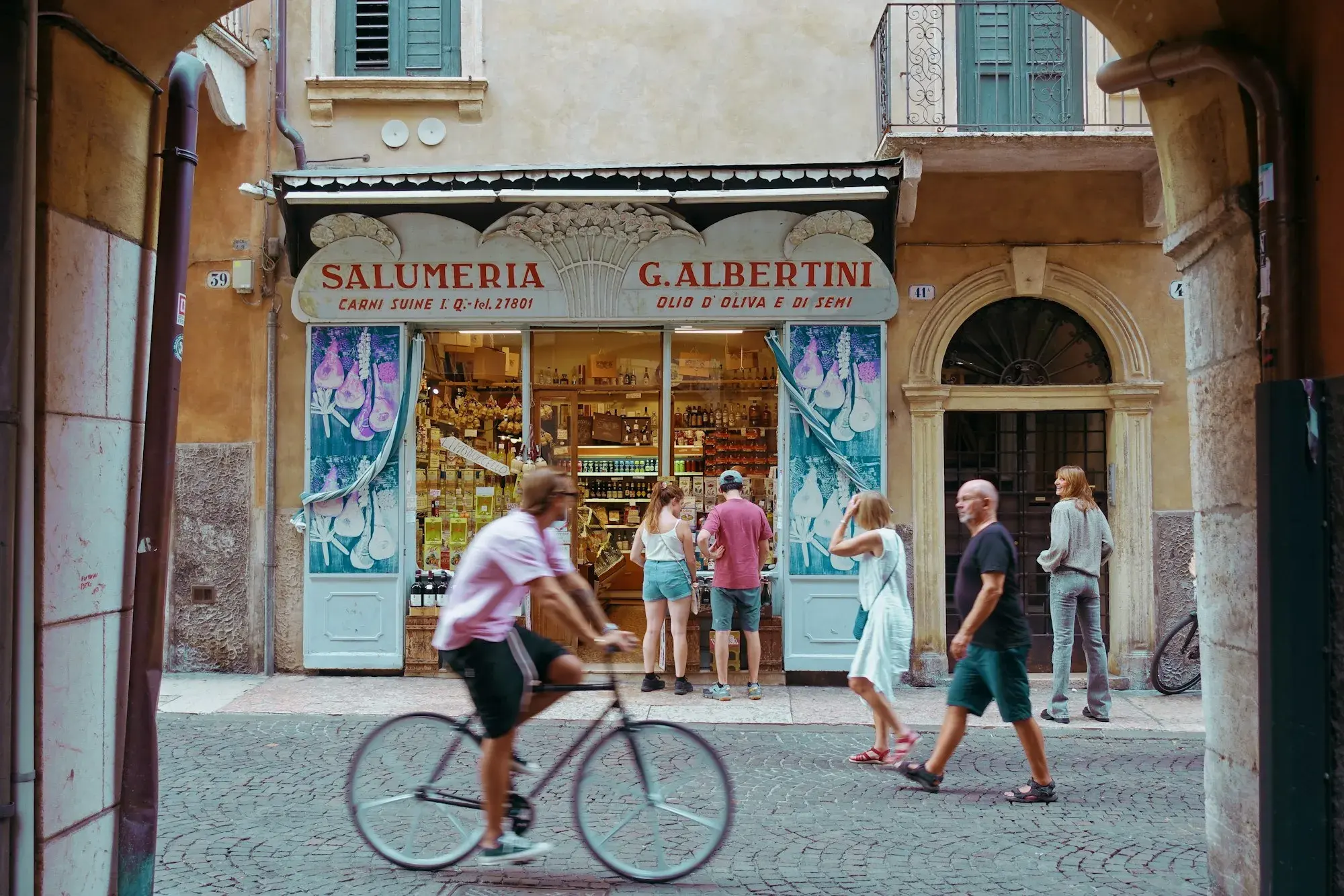 A cyclist and pedestrians passing a traditional delicatessen with a painted sign on a street in Verona