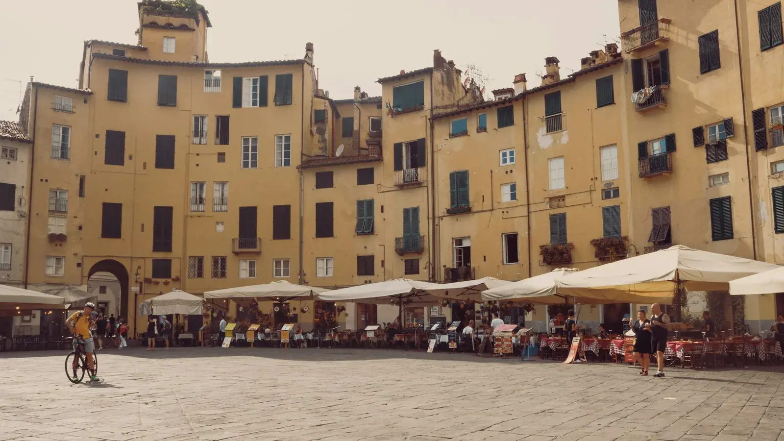 Cyclists and pedestrians crossing a sunlit piazza in central Lucca, framed by historic stone arcades