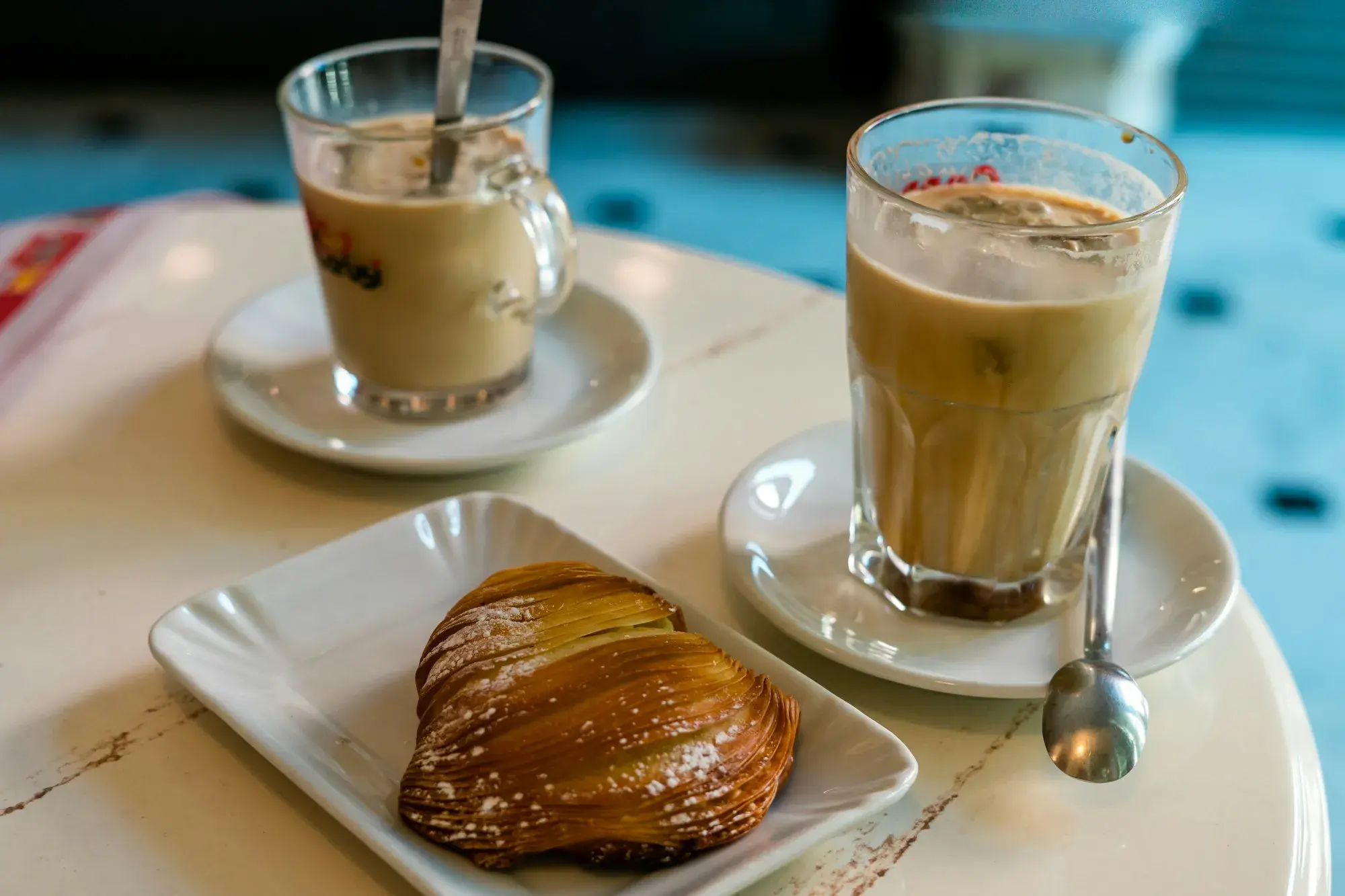 Two glasses of iced coffee on saucers beside a crisp, shell-shaped sfogliatella pastry on a café table