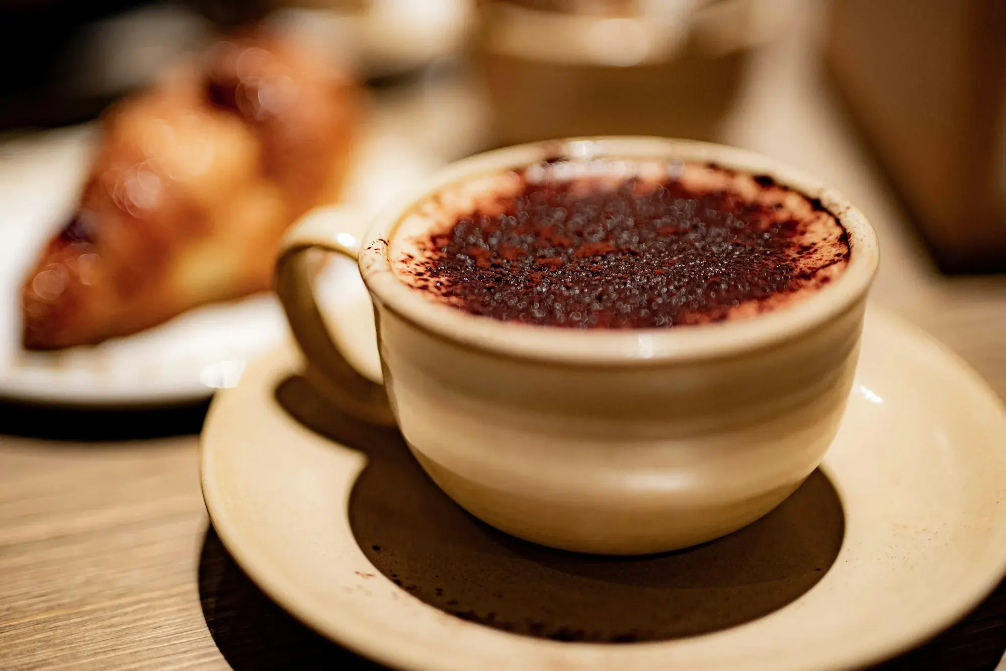 A cappuccino dusted with cocoa in a cream-coloured cup, with a sugared brioche on a plate behind it