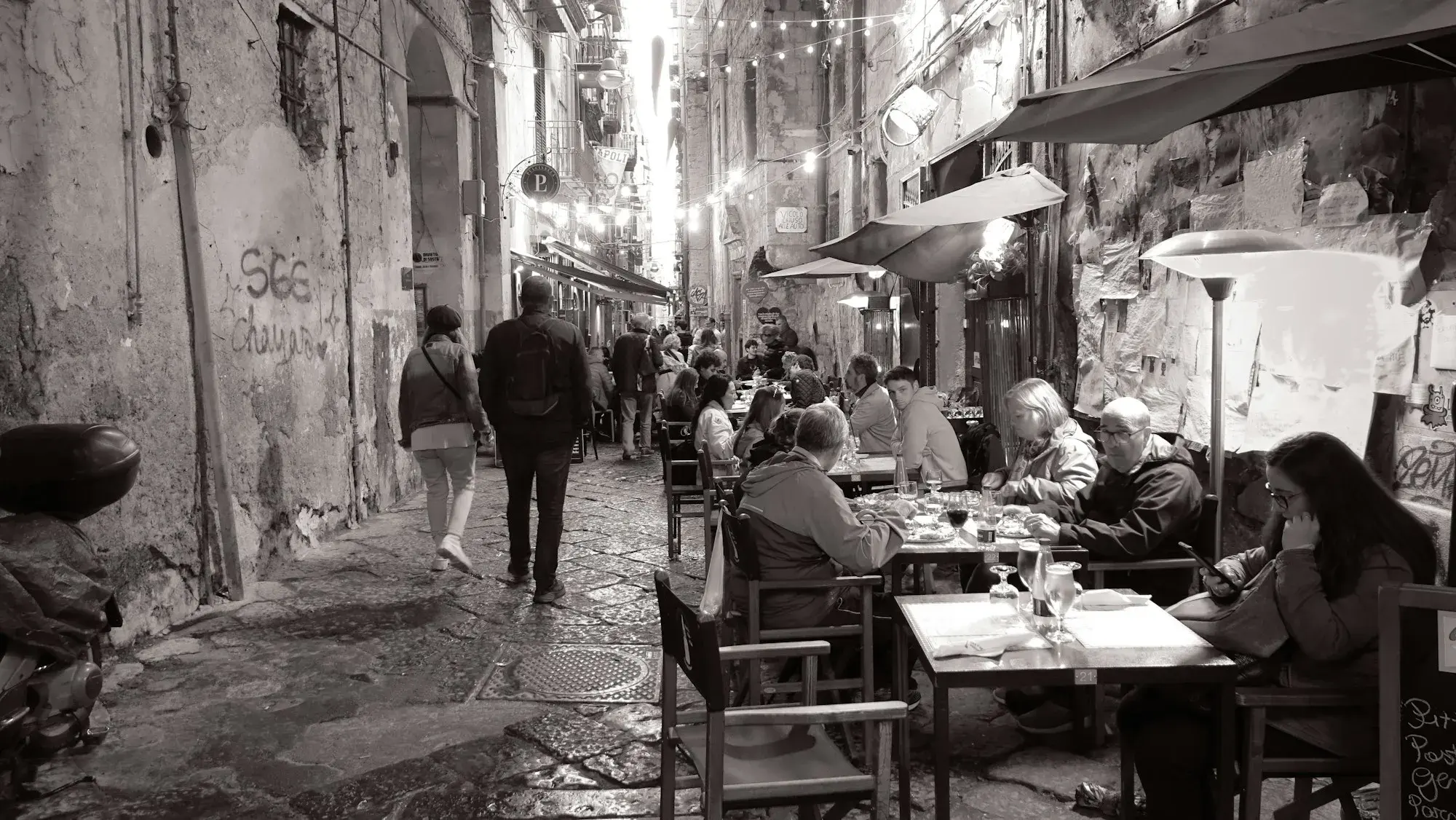 Black-and-white photo of people at café tables along a narrow street in central Naples, with strings of lights overhead