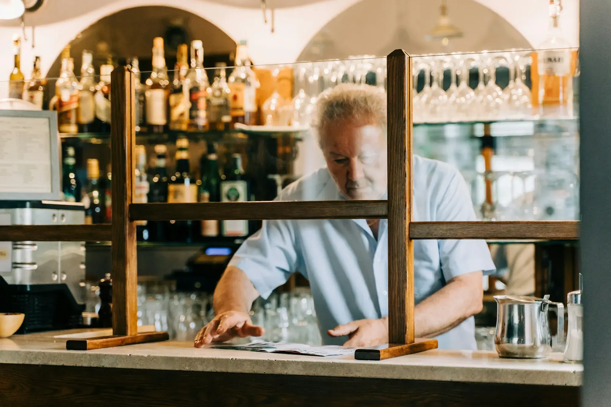 A barman in a light-blue shirt working behind the wooden counter of a village bar, with bottles on shelves and a clear screen along the counter