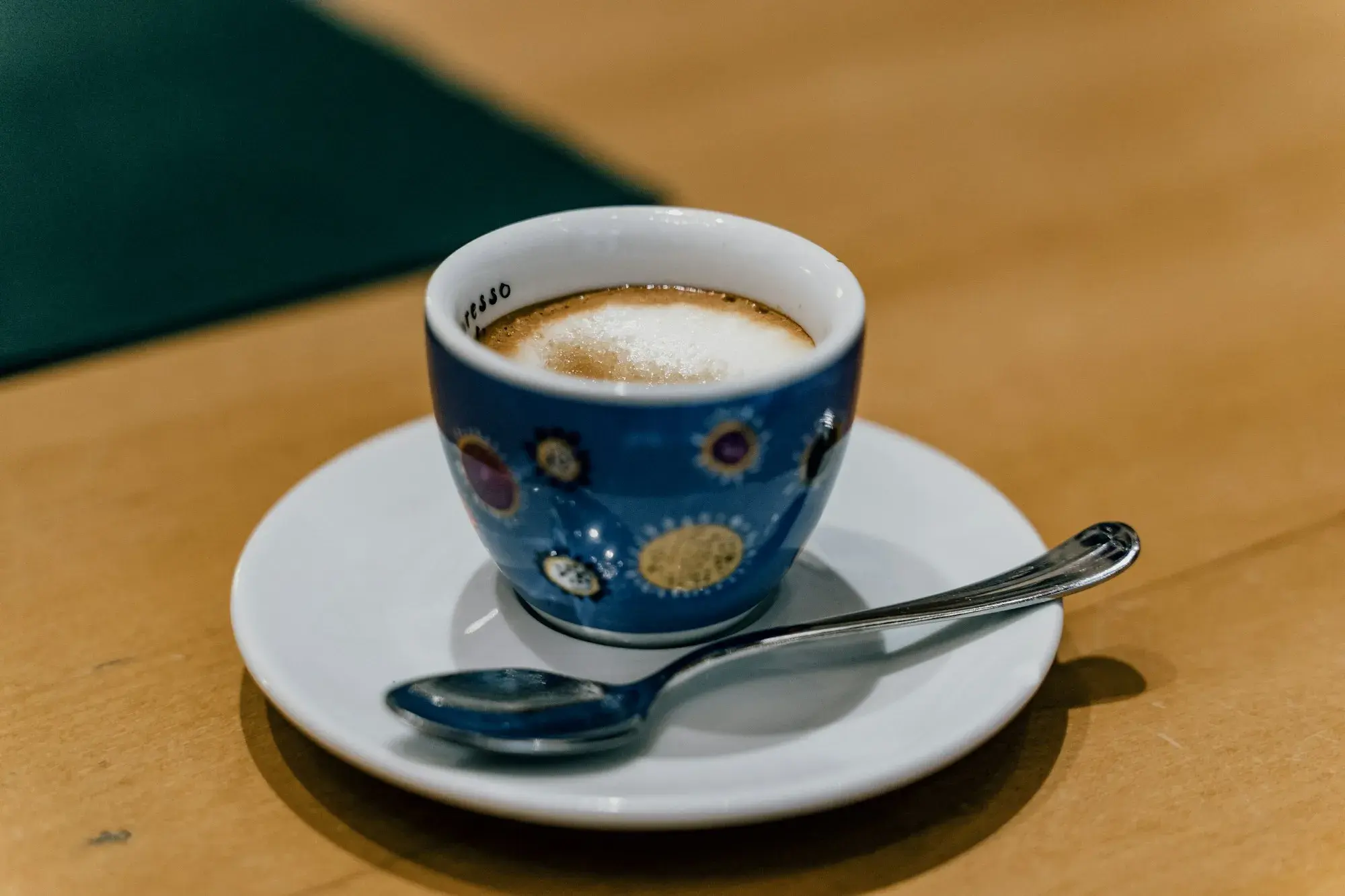 A small espresso with a light layer of foam in a patterned blue cup on a white saucer with a spoon, on a wooden bar table in Rome
