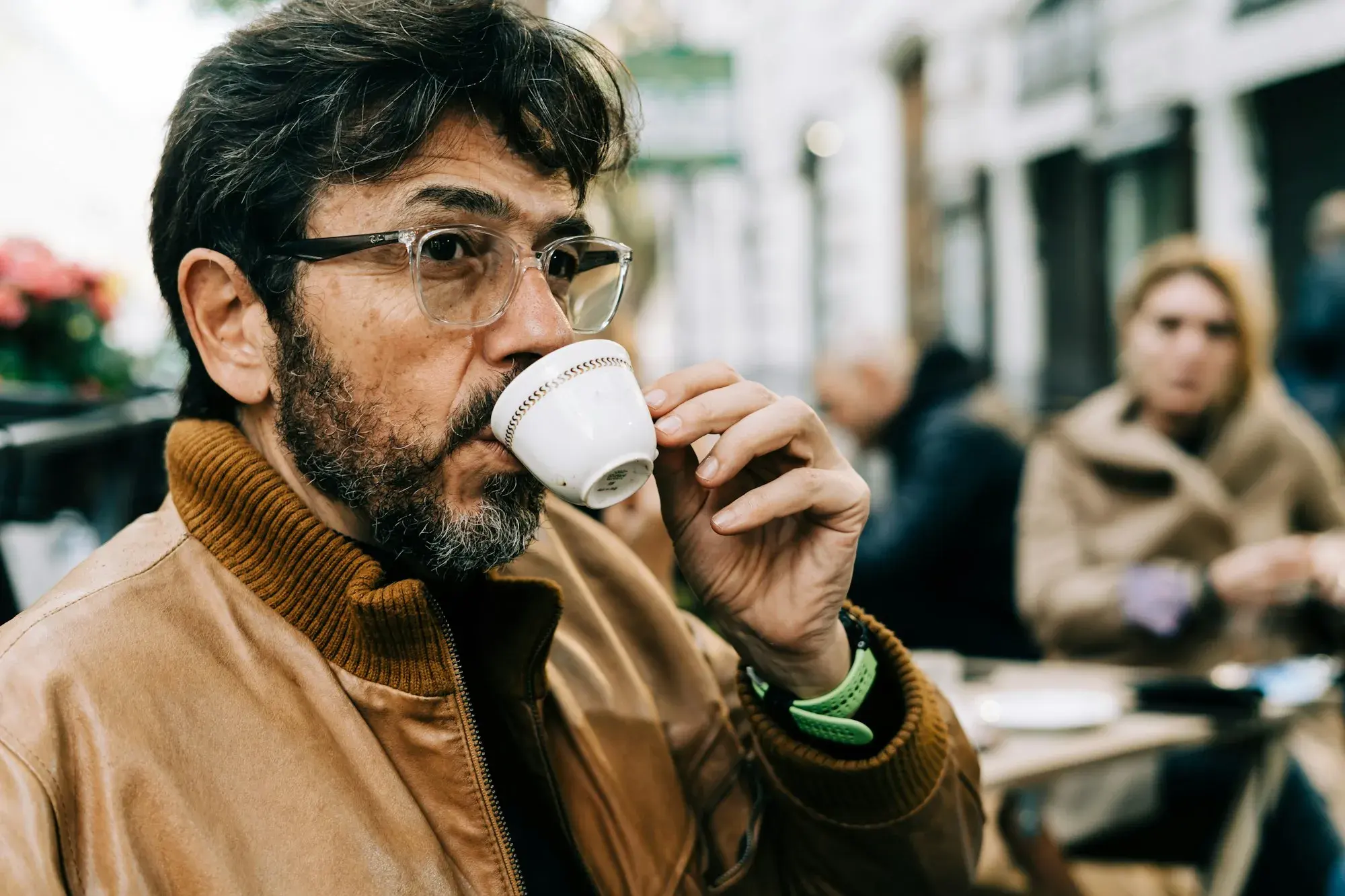A man with glasses and a beard in a tan leather jacket sipping an espresso at an outdoor café table in Rome