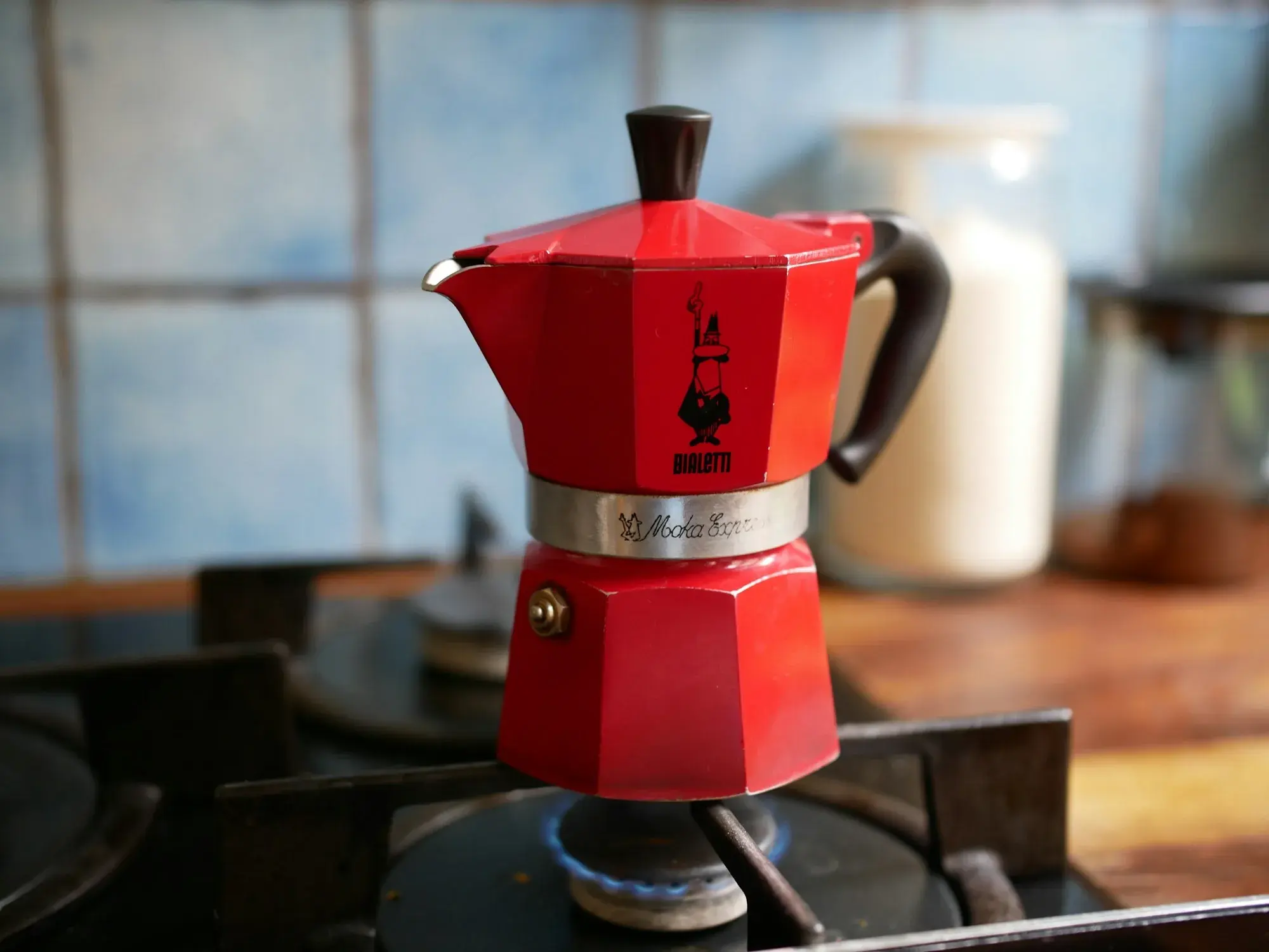 A small red Bialetti moka pot on a gas ring in a home kitchen in Rome, with a blue flame beneath