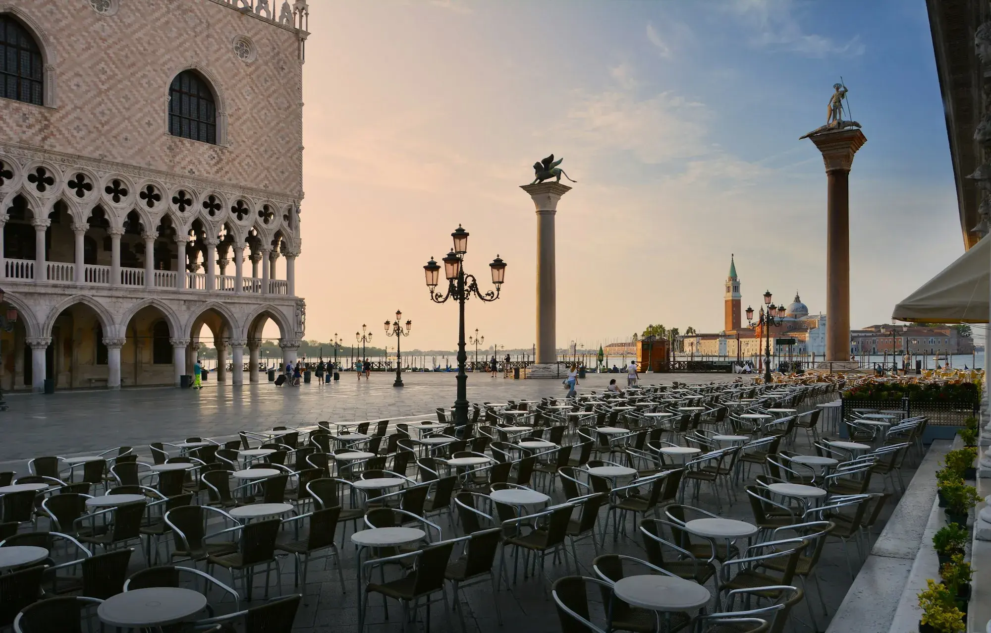 Rows of empty café tables and chairs on the Piazzetta di San Marco at dawn, beside the Doge's Palace and its two columns
