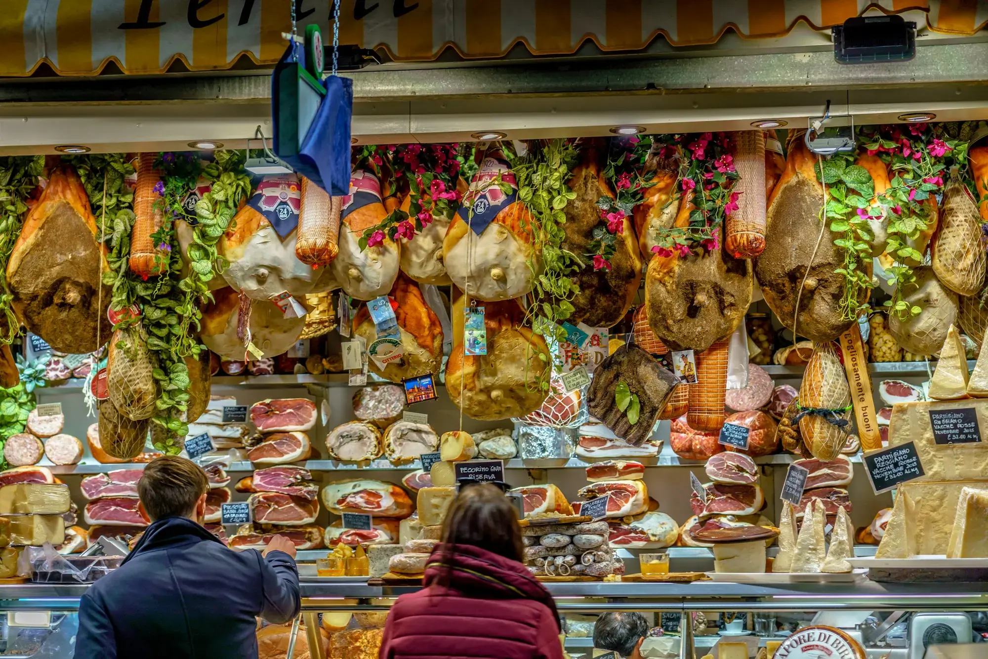 Clienti a un banco del Mercato Centrale di Firenze sotto prosciutti, salami e formaggi appesi