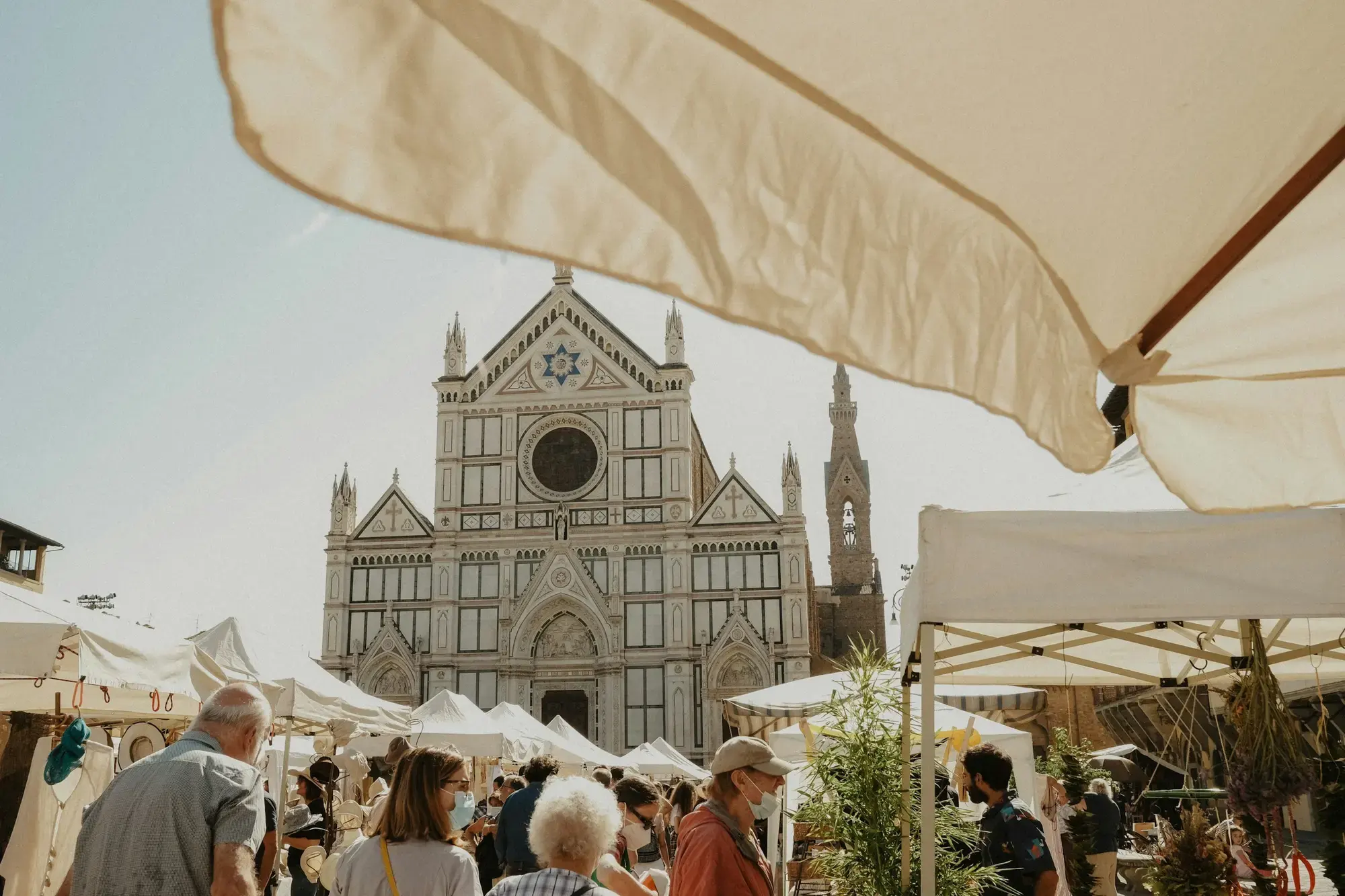 Persone tra i banchi coperti da tendoni bianchi in piazza Santa Croce a Firenze, con la facciata in marmo della basilica sullo sfondo