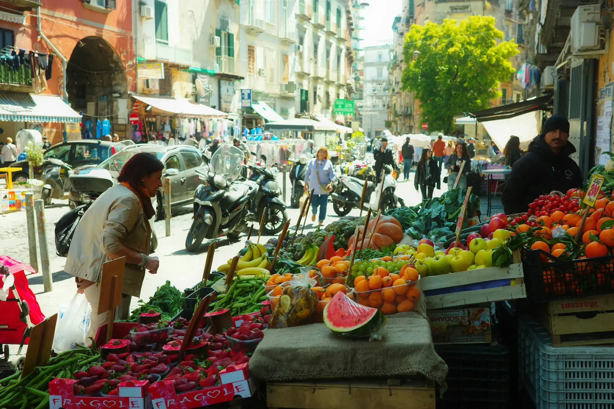 A woman shopping at a fruit and vegetable stall piled with oranges, melons and greens on a busy street market in Naples, with scooters and shopfronts behind