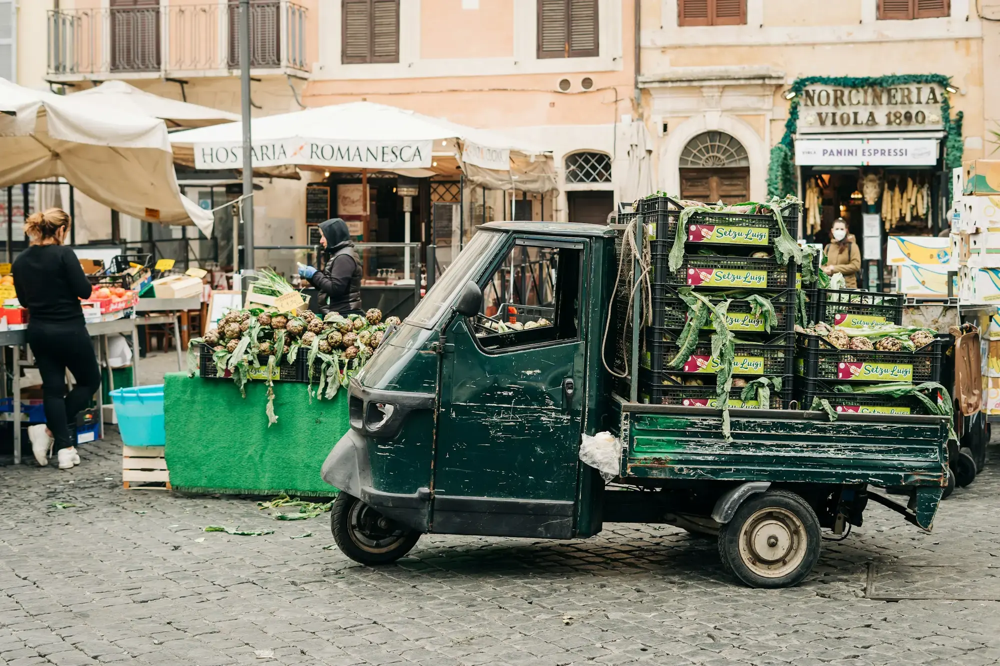 Un'Ape carica di cassette di carciofi tra i banchi di Campo de' Fiori a Roma, con una trattoria e una norcineria sullo sfondo