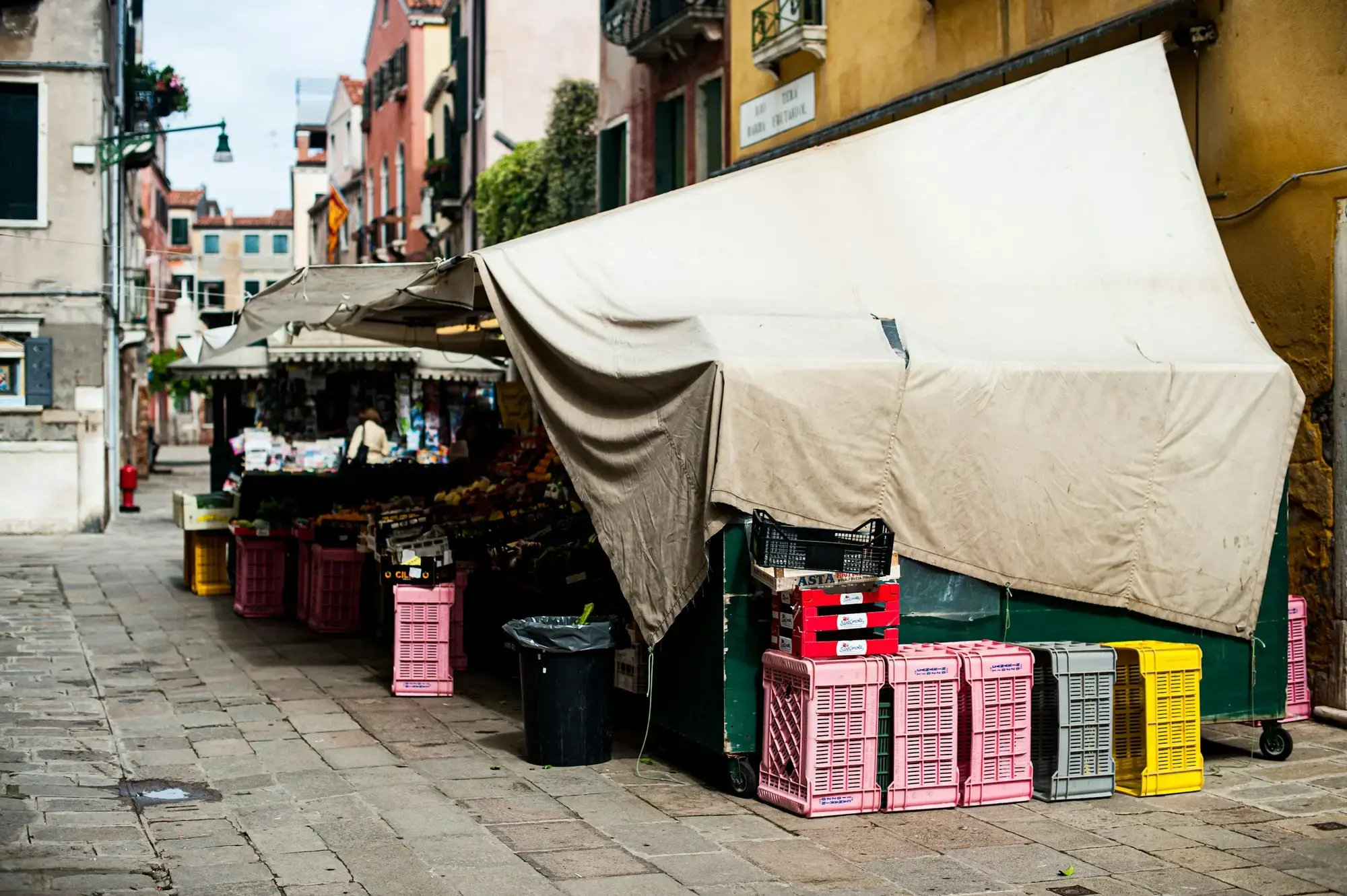 Un banco di frutta e verdura in una calle veneziana, coperto a metà da un telo, con cassette impilate sul selciato