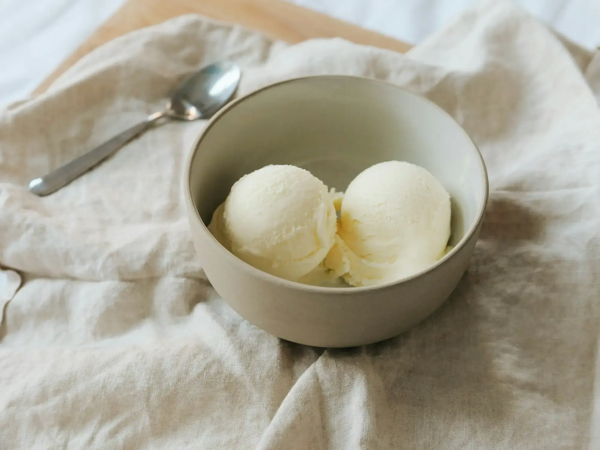 Two scoops of white gelato in a pale ceramic bowl on a linen cloth, with a spoon beside it
