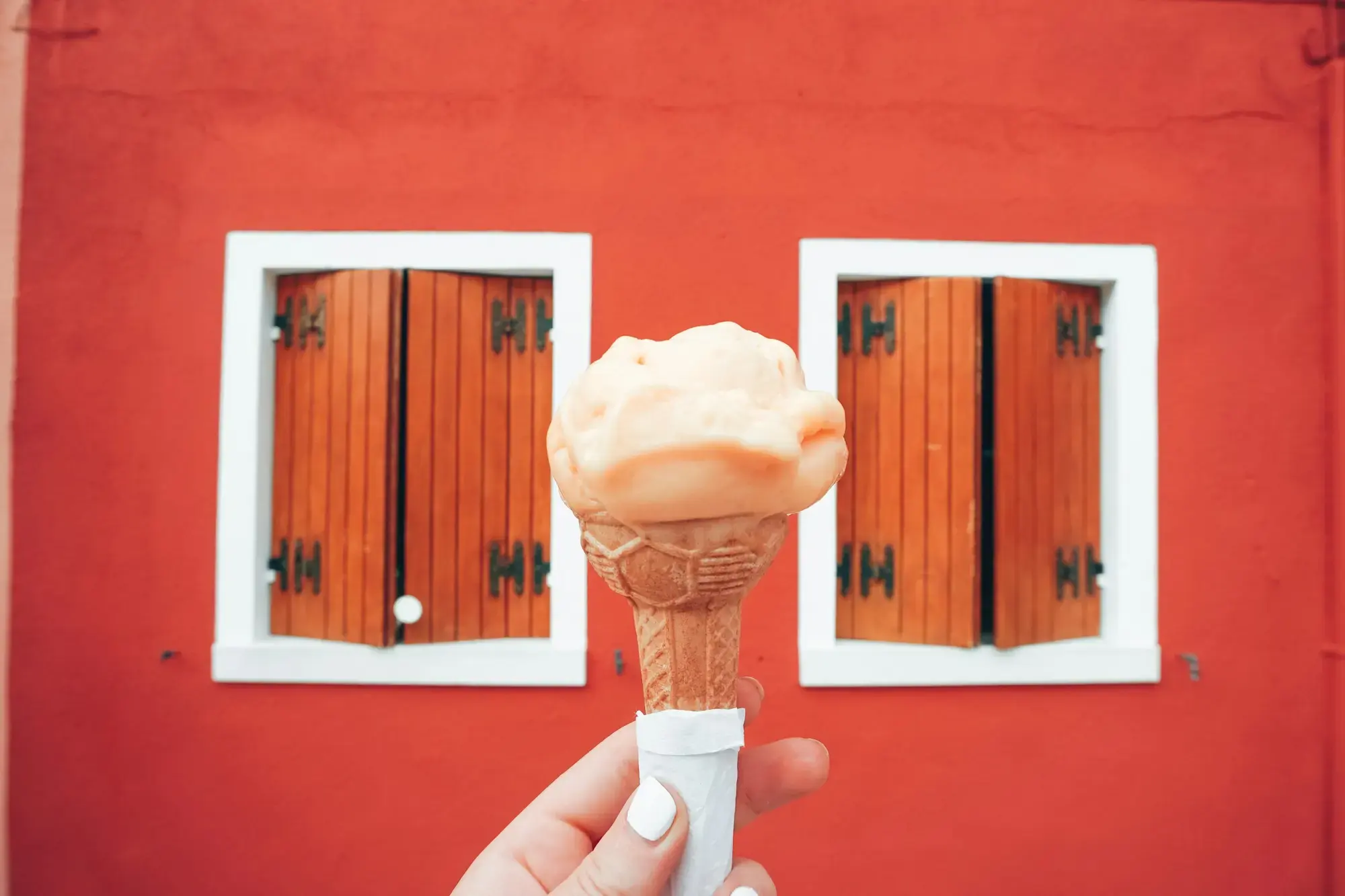 A hand holding up a cone of pale gelato in front of a red-painted wall between two windows with wooden shutters