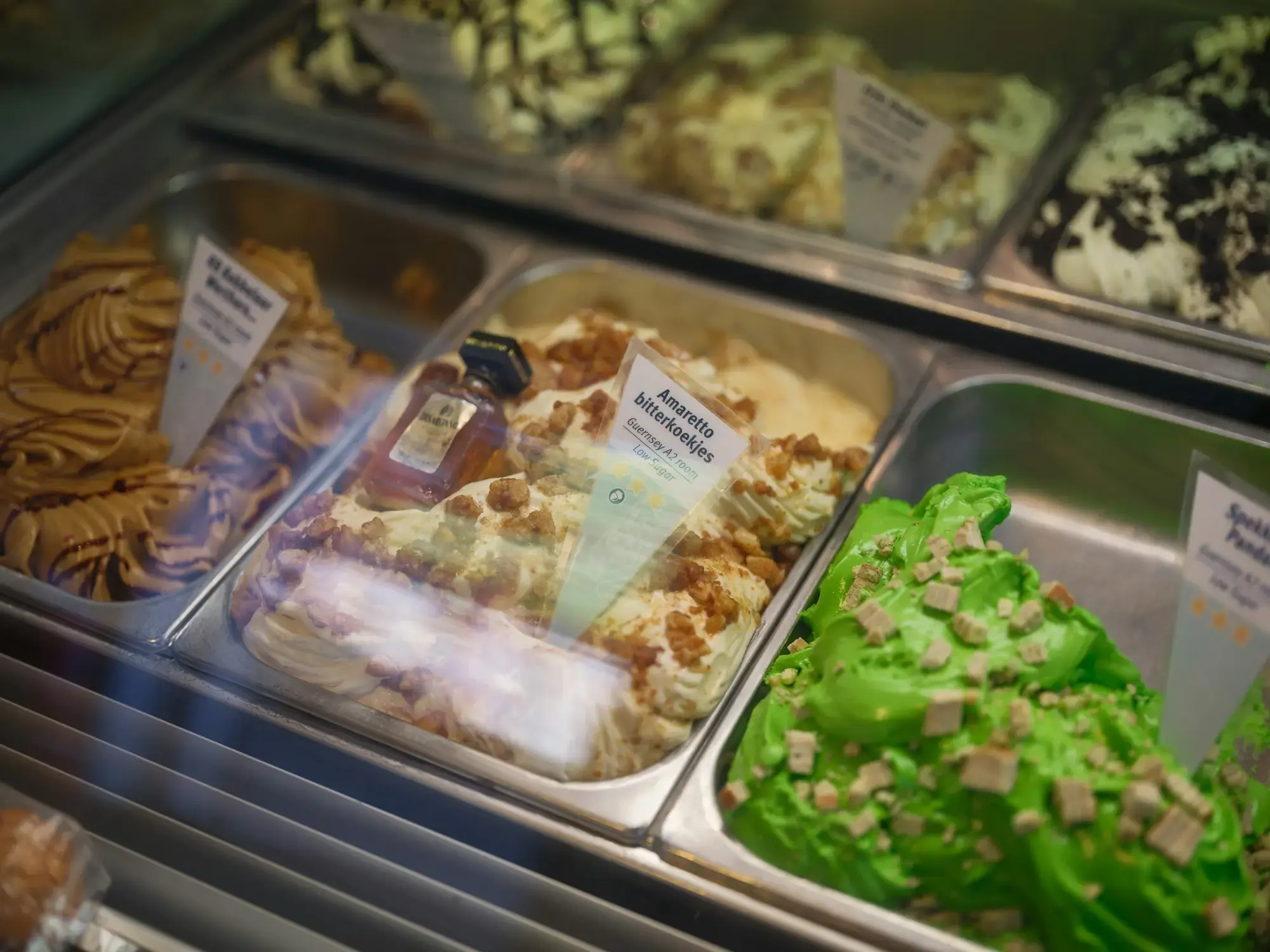 A gelato display case with a vivid green flavour beside paler nut, biscuit and cream flavours in steel trays, each with a label