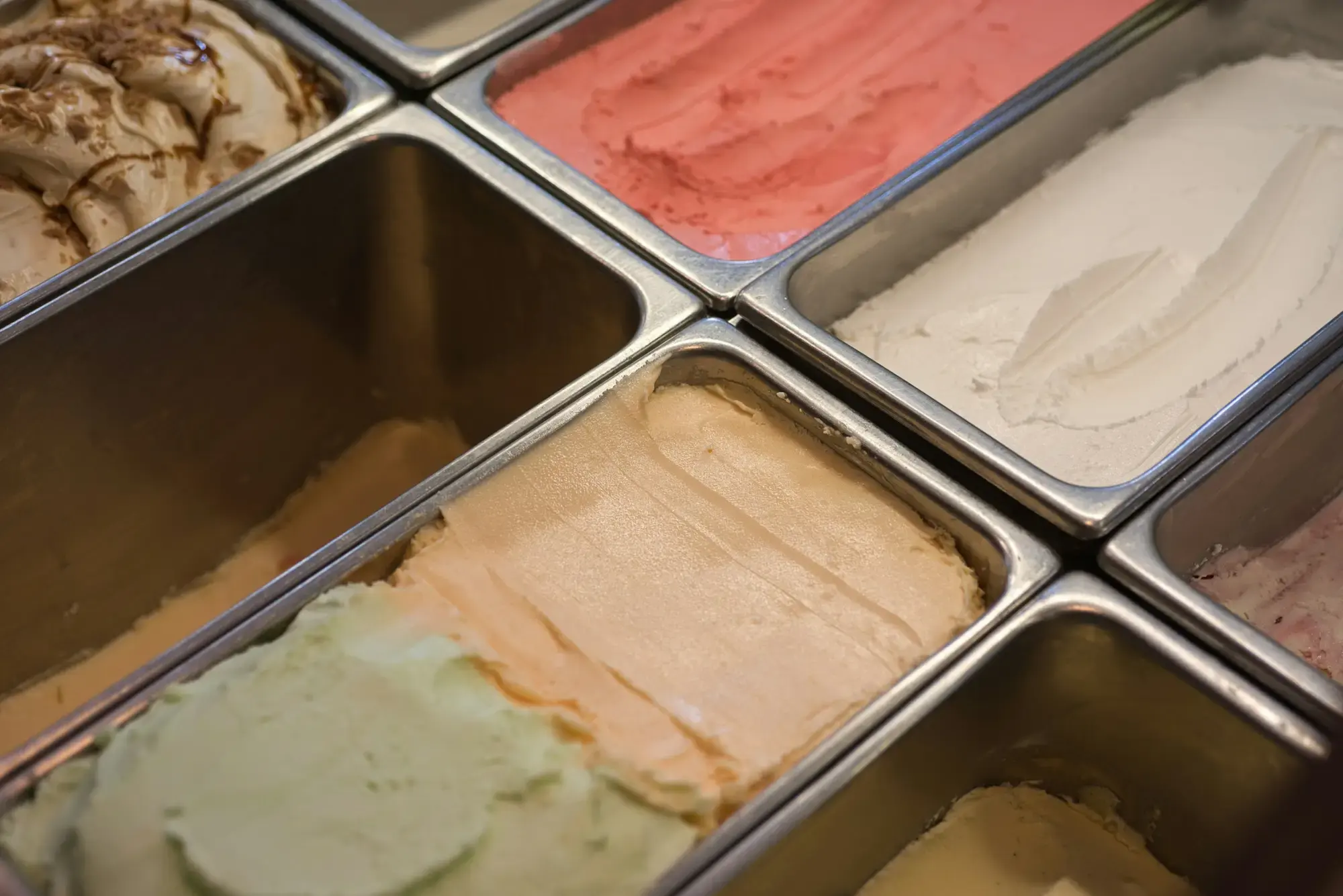 Stainless-steel trays of gelato in a display case, with pale nut, cream and pink fruit flavours smoothed flat