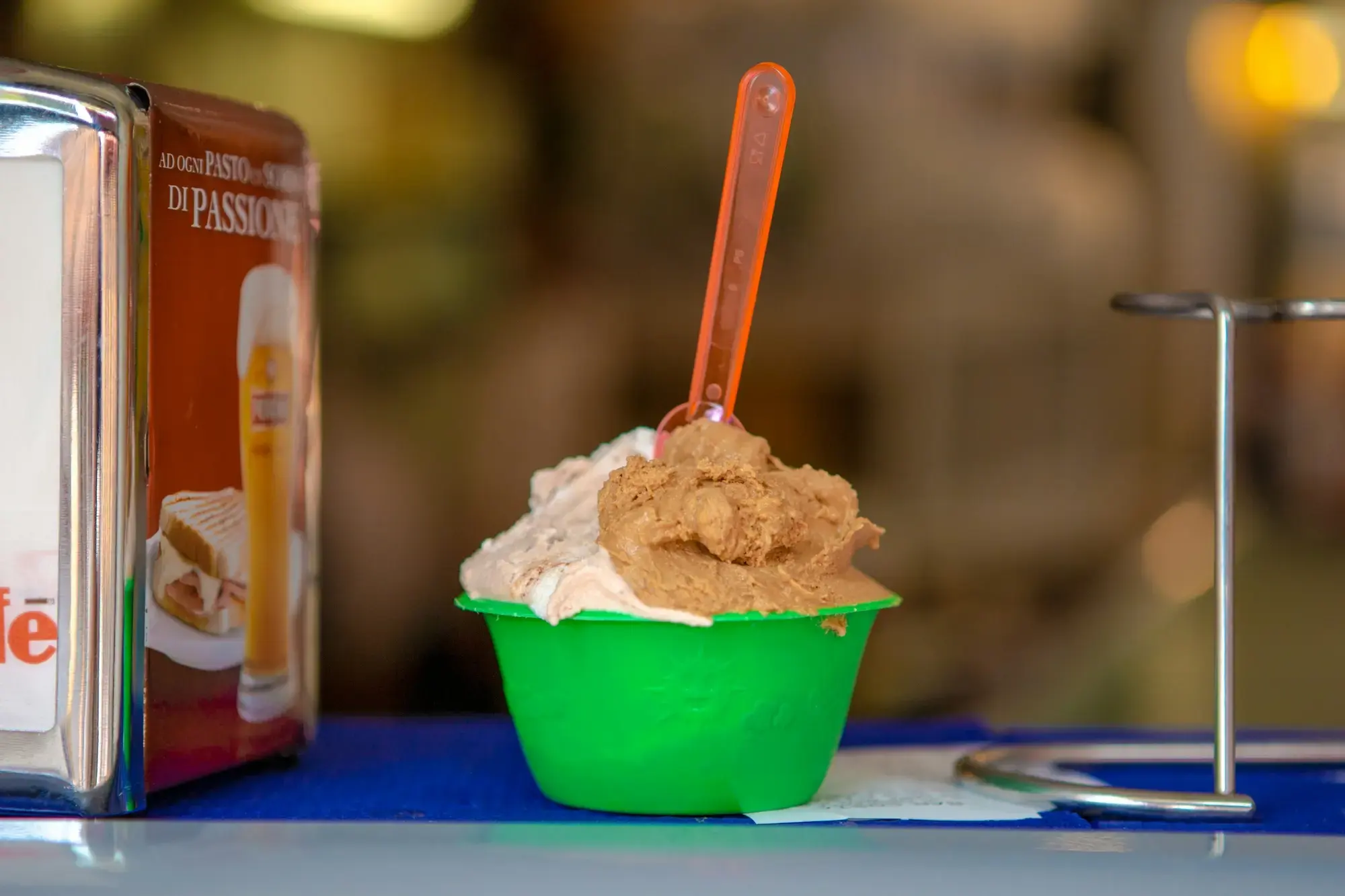 A green paper cup of gelato with a small orange plastic spoon, on a counter in Pisa