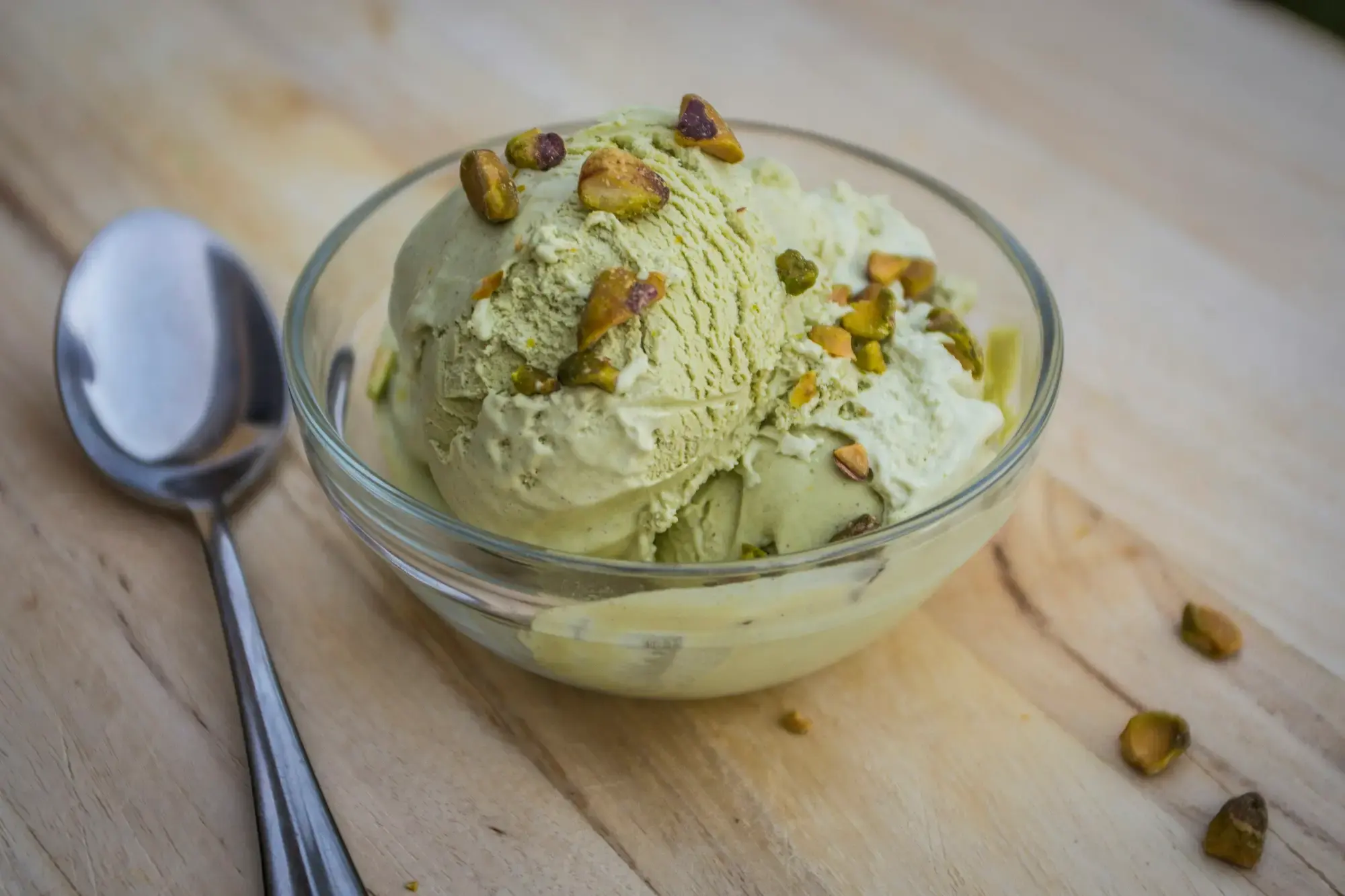 A scoop of pale green pistachio gelato in a glass bowl, scattered with chopped pistachios, with a spoon beside it