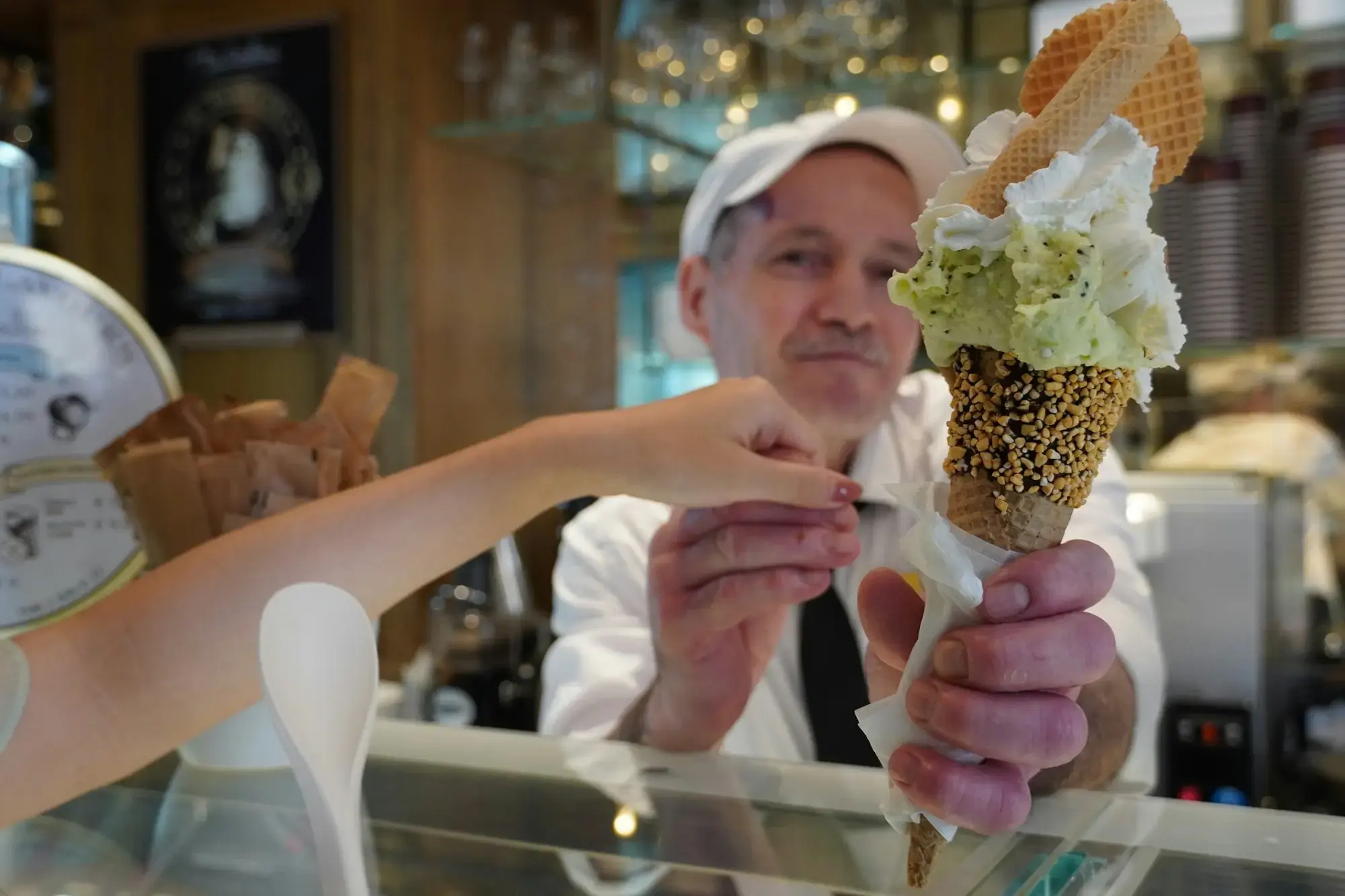 A gelato maker in a white cap holding out a cone of pale green and white gelato topped with whipped cream across the counter of a gelateria in Rome