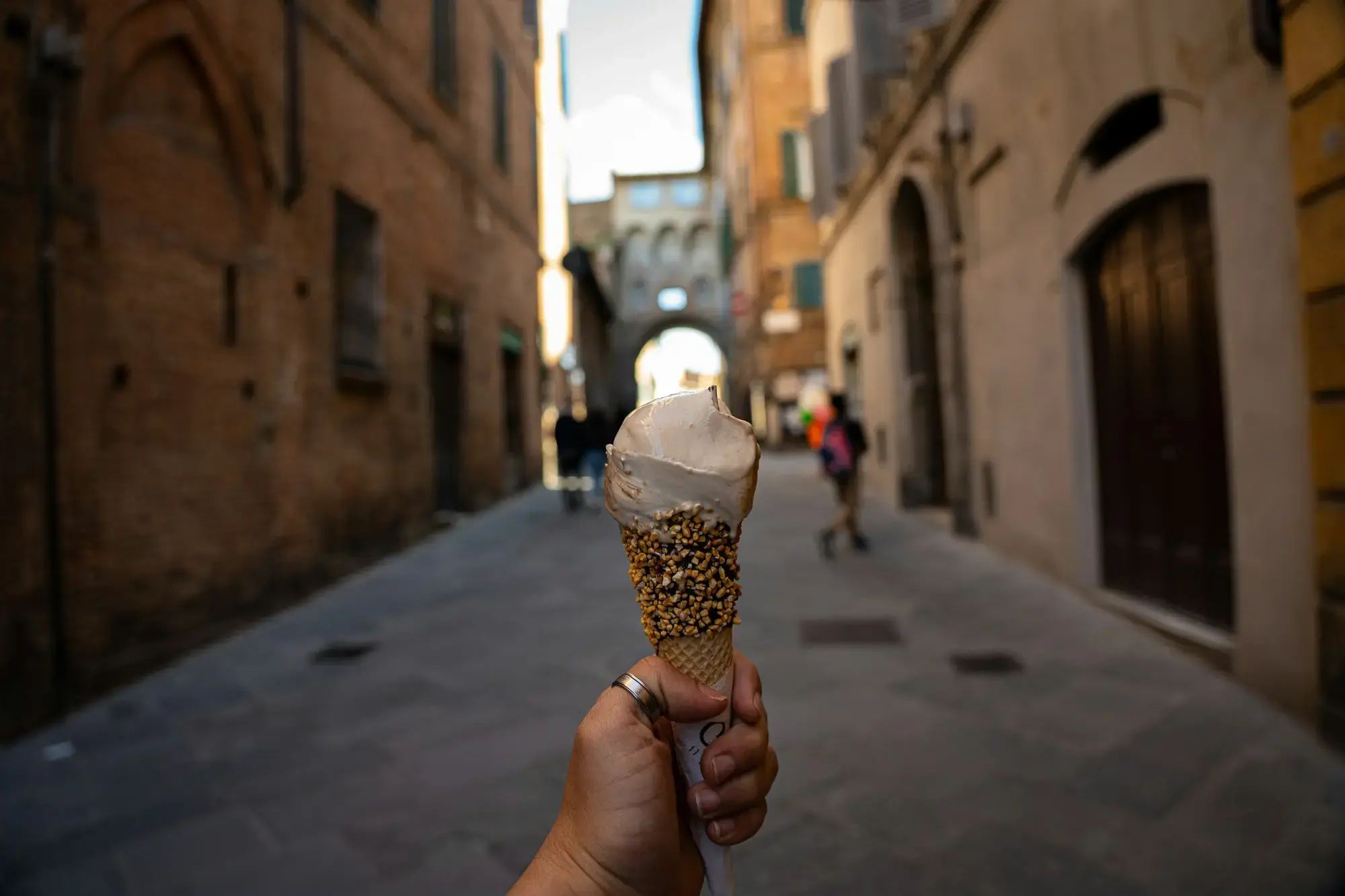 A hand holding a cone of gelato up in a narrow, shaded street of old stone buildings in Siena