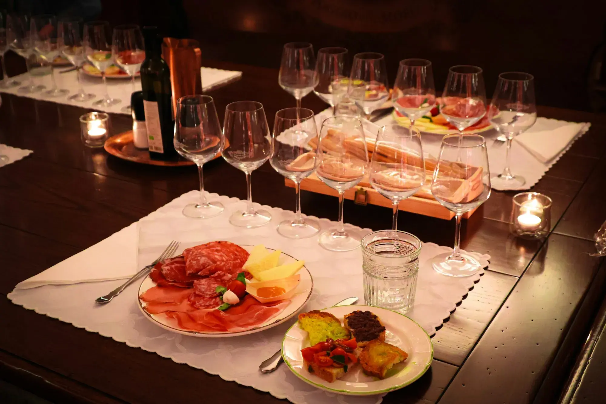 A candlelit table set for a wine tasting, with rows of glasses, a bottle, and plates of cured meats and bruschetta