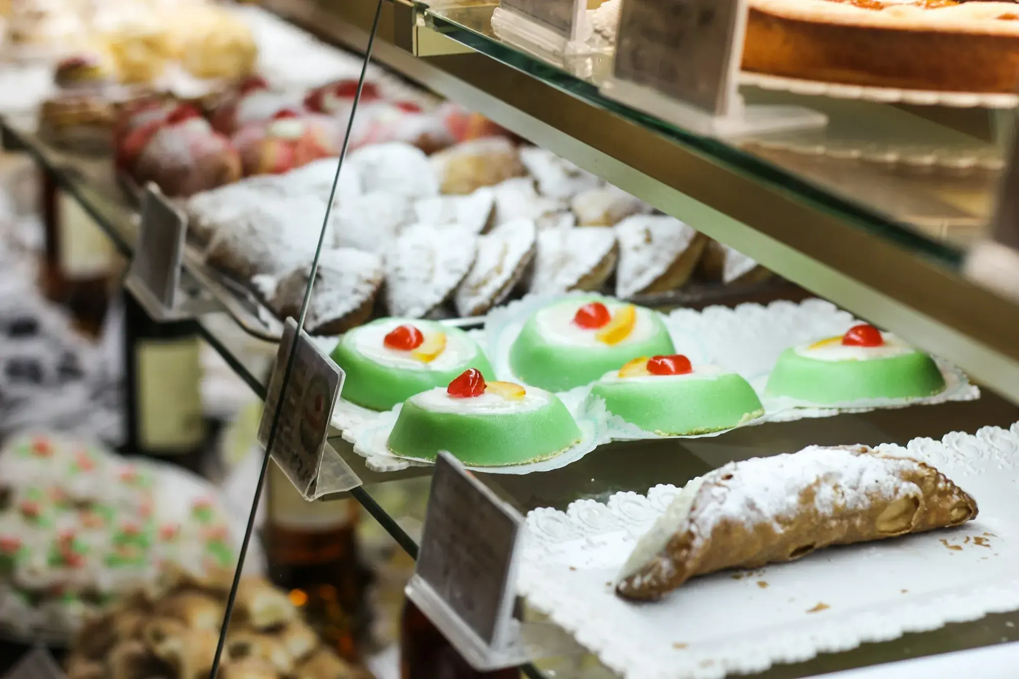 A pastry counter in Erice with green iced cassatine, a sugar-dusted cannolo and trays of almond biscuits