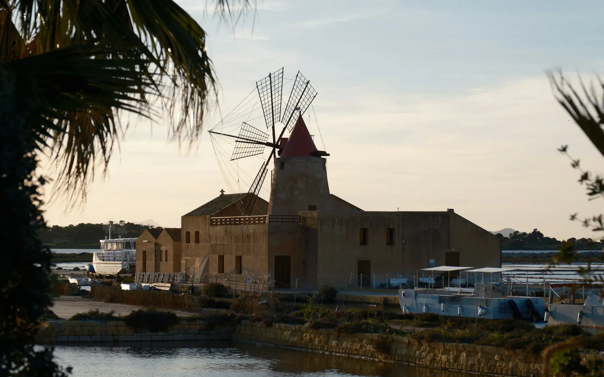 A stone windmill and low salt-works buildings beside a salt pan near Marsala, framed by palm leaves in evening light