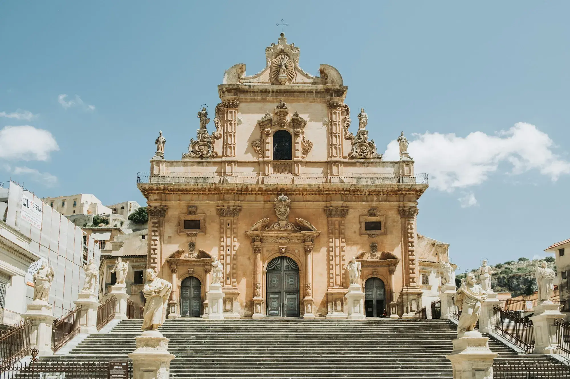 The Baroque façade of a church in Modica, with statues lining a wide flight of steps under a blue sky