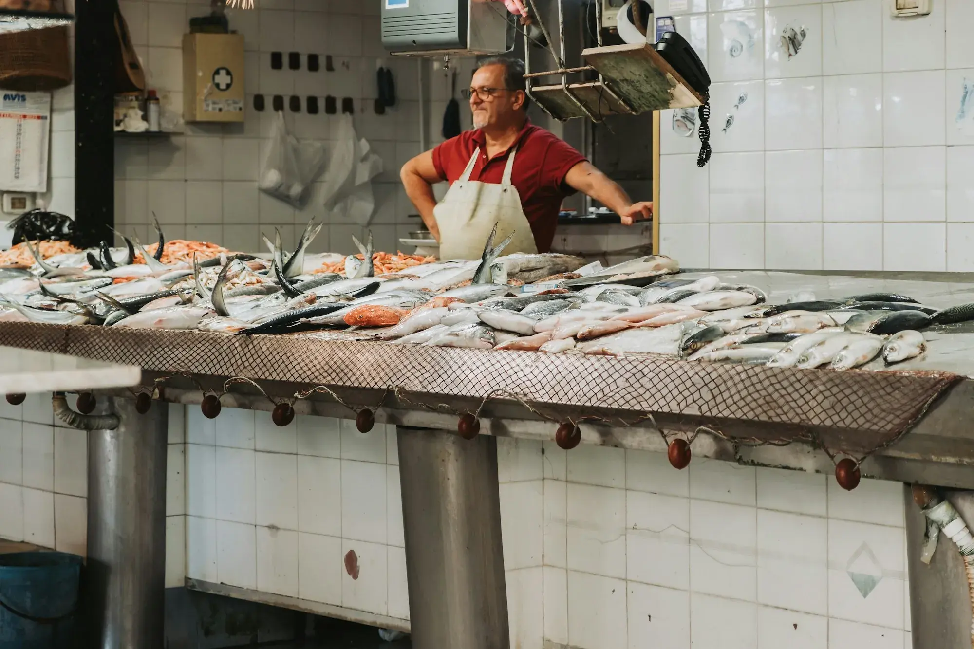 A fishmonger in a red shirt and white apron behind a long tiled counter of fresh fish at the market in Ortigia, Syracuse