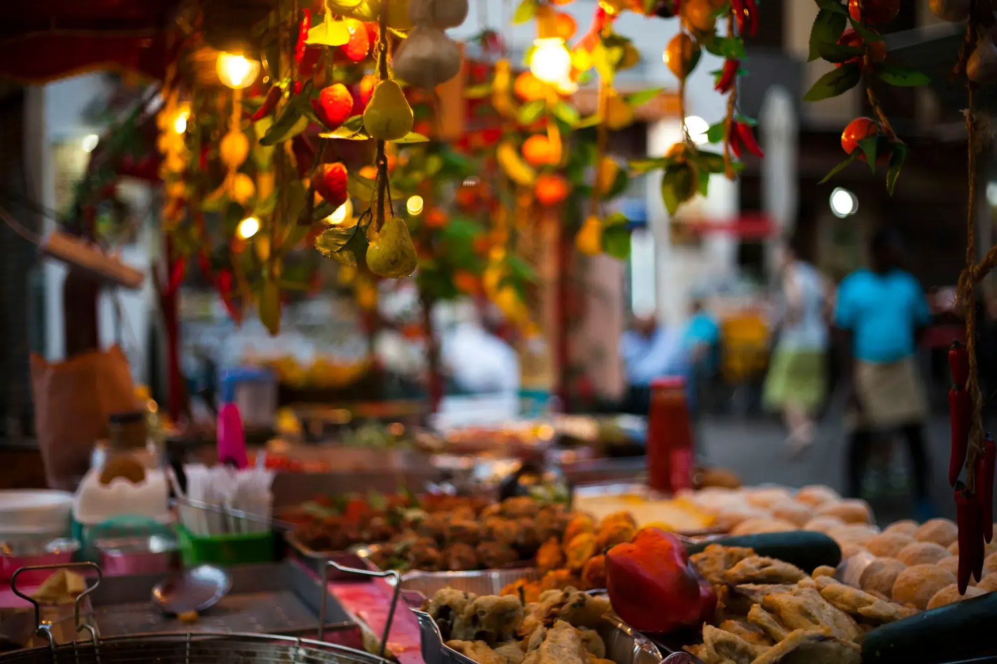 Market stalls in Palermo at dusk, with fried snacks in the foreground and strings of peppers and lights hanging above