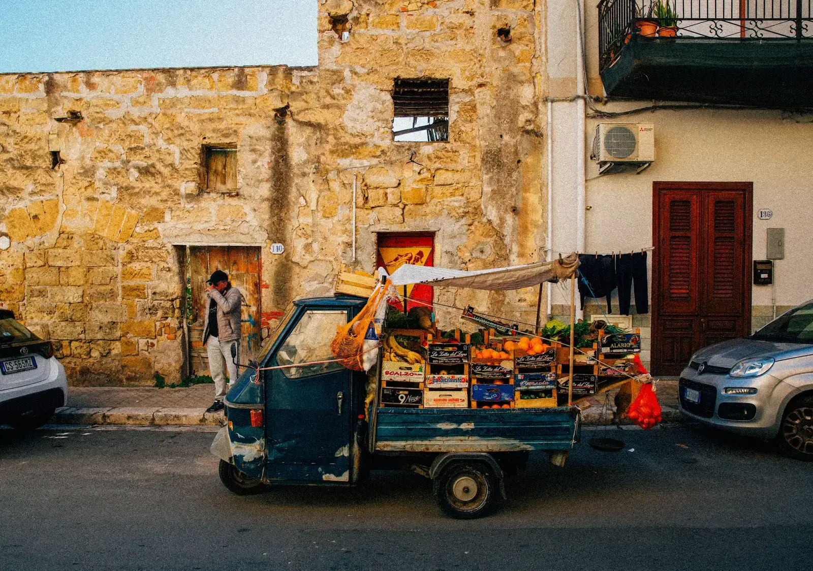 A three-wheeled Piaggio Ape loaded with crates of oranges and produce parked on a street in Palermo beside a stone house