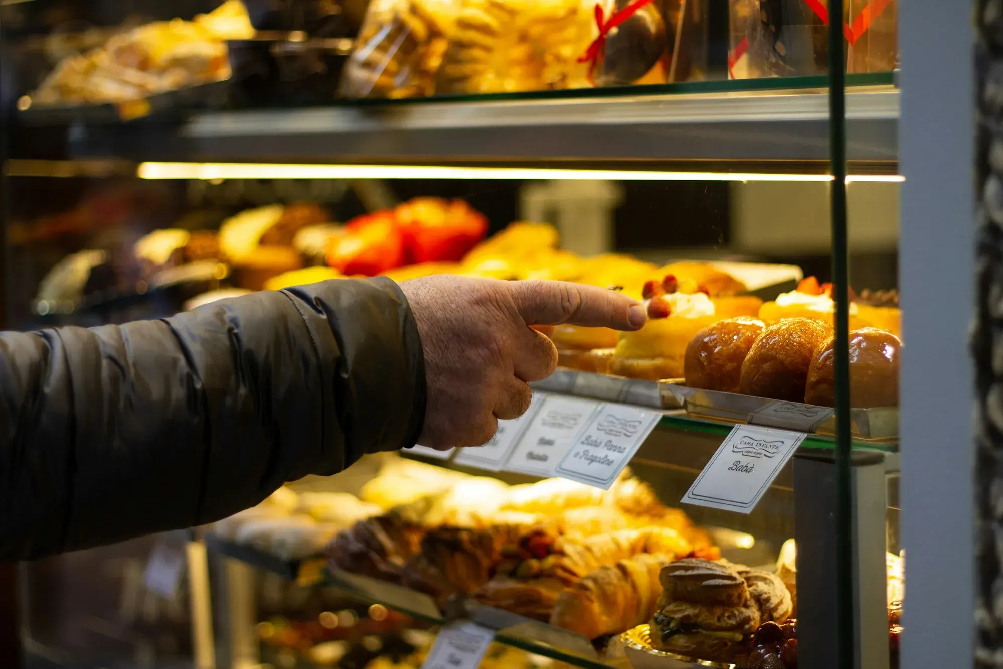 A customer points at trays of glazed pastries in a lit glass display case at a pastry shop in Naples