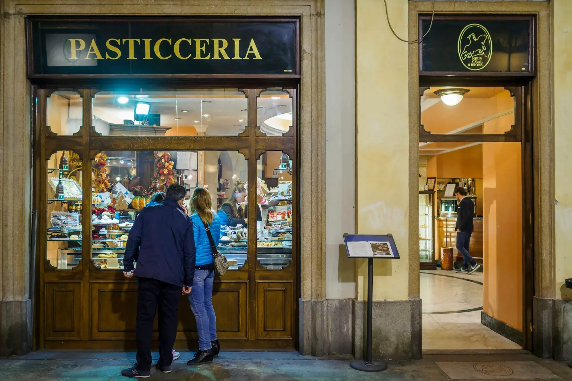 Two people looking into the lit window of a traditional pasticceria with a dark wooden shopfront in central Turin