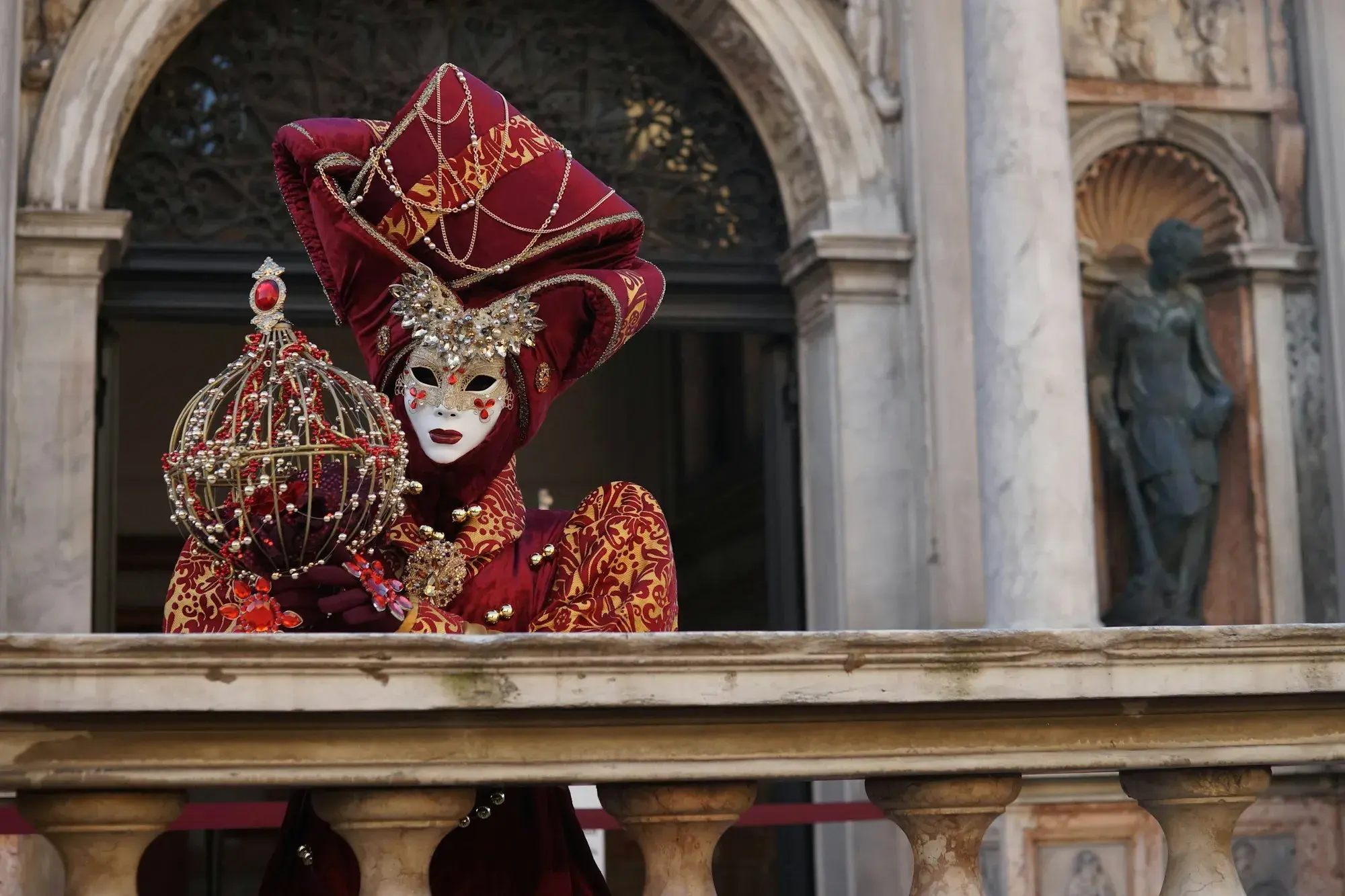 A person in an elaborate red and gold Venetian Carnival costume and white mask on a stone balcony