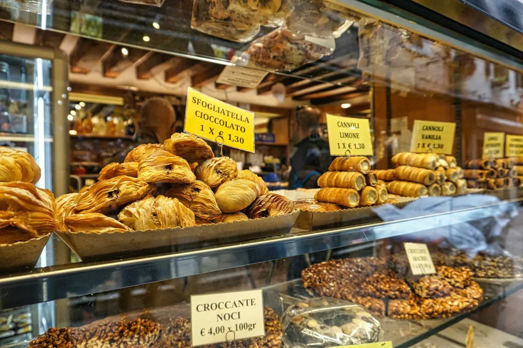 A pastry-shop counter in Venice with trays of puff-pastry sfogliatelle, cream-filled cannoli and nut brittle under yellow price signs