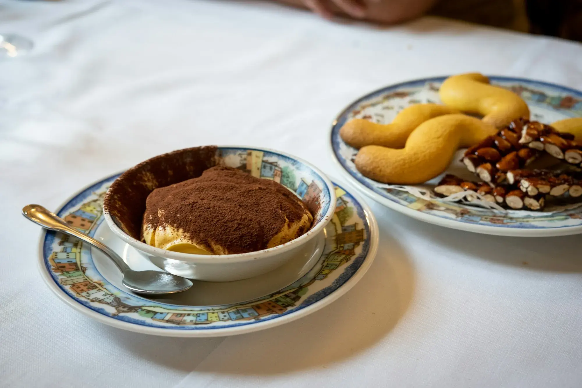 A cup of tiramisù dusted with cocoa beside a plate of biscuits and nut brittle on a white tablecloth in Venice
