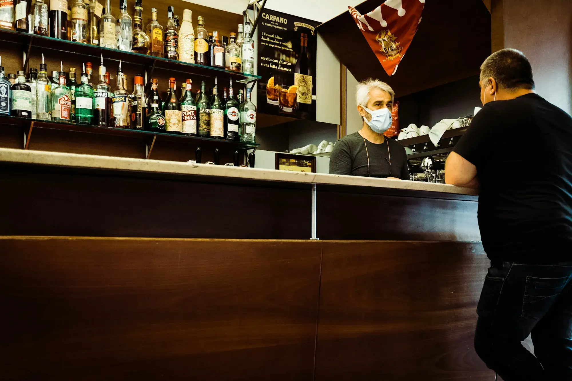 A bartender chatting with a customer across the counter of a small bar in San Quirico d'Orcia, Tuscany