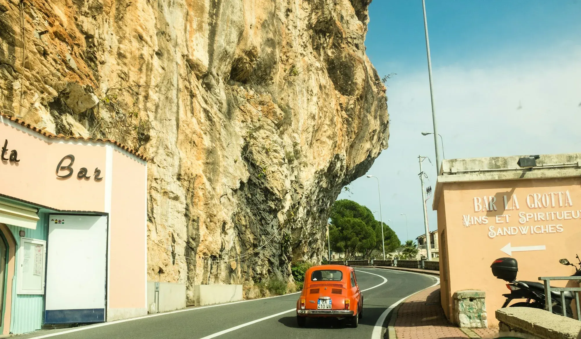 A small red car on a narrow road between rocky cliffs and buildings on the Ligurian coast near Grimaldi